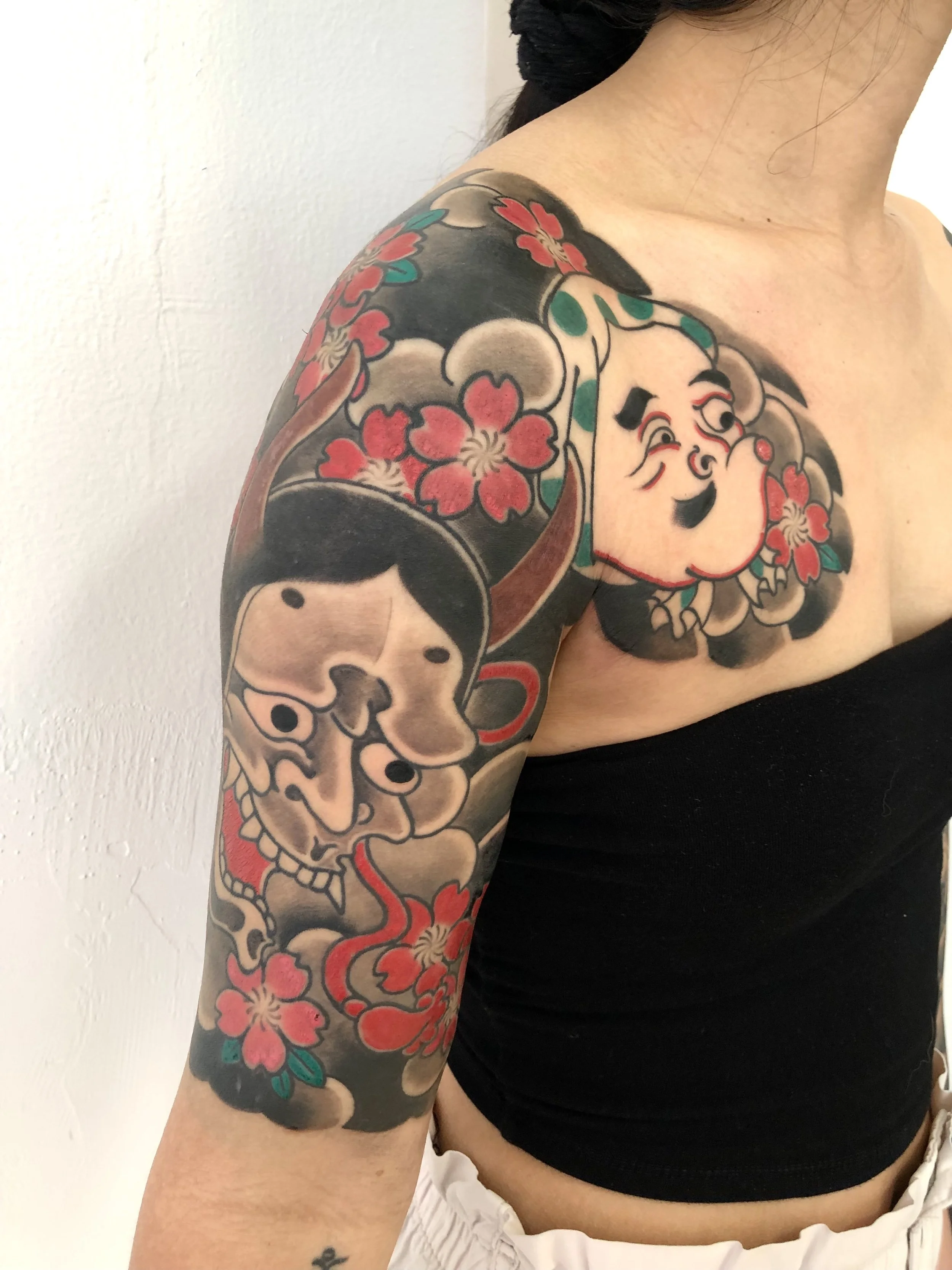Colorful tattoo on woman's shoulder and upper arm featuring a traditional Japanese style design with a smiling face, a dog, and pink cherry blossoms.