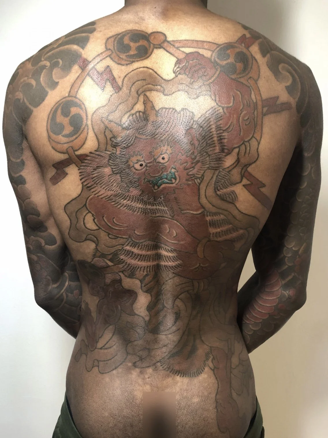 A person's back with extensive traditional-style tattoos, including a large central Japanese demon face, dragons, clouds, and interwoven patterns.