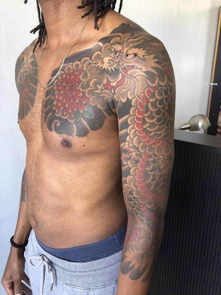 A man with shoulder-length dreadlocks and a gold necklace showing his upper body tattoos, including a large dragon on his shoulder and arm, and a red flower on his chest.