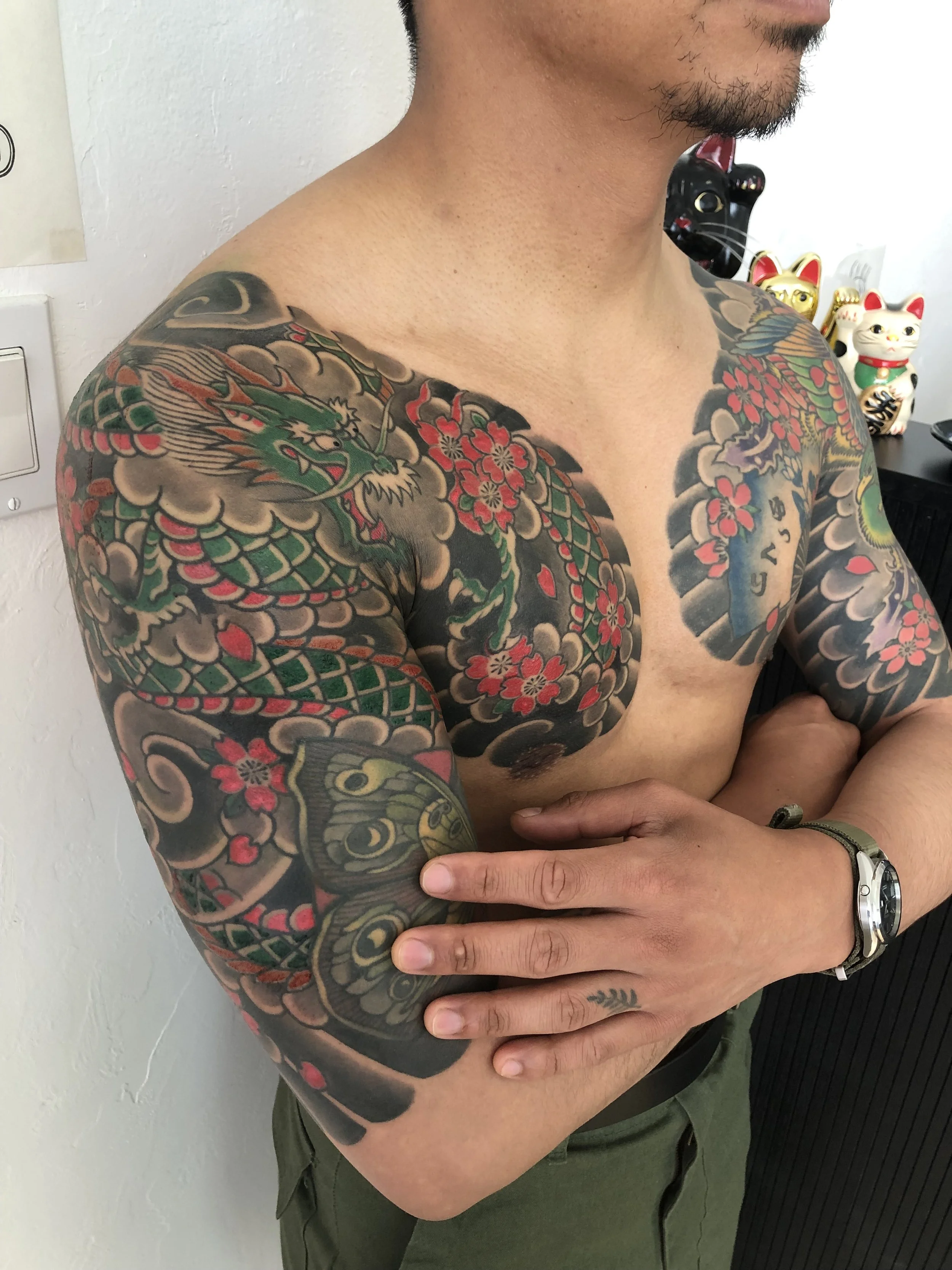A shirtless man with a large colorful tattoo of a dragon, cherry blossoms, and a tiger on his arms and chest, standing with arms crossed against a white wall with Japanese figurines in the background.