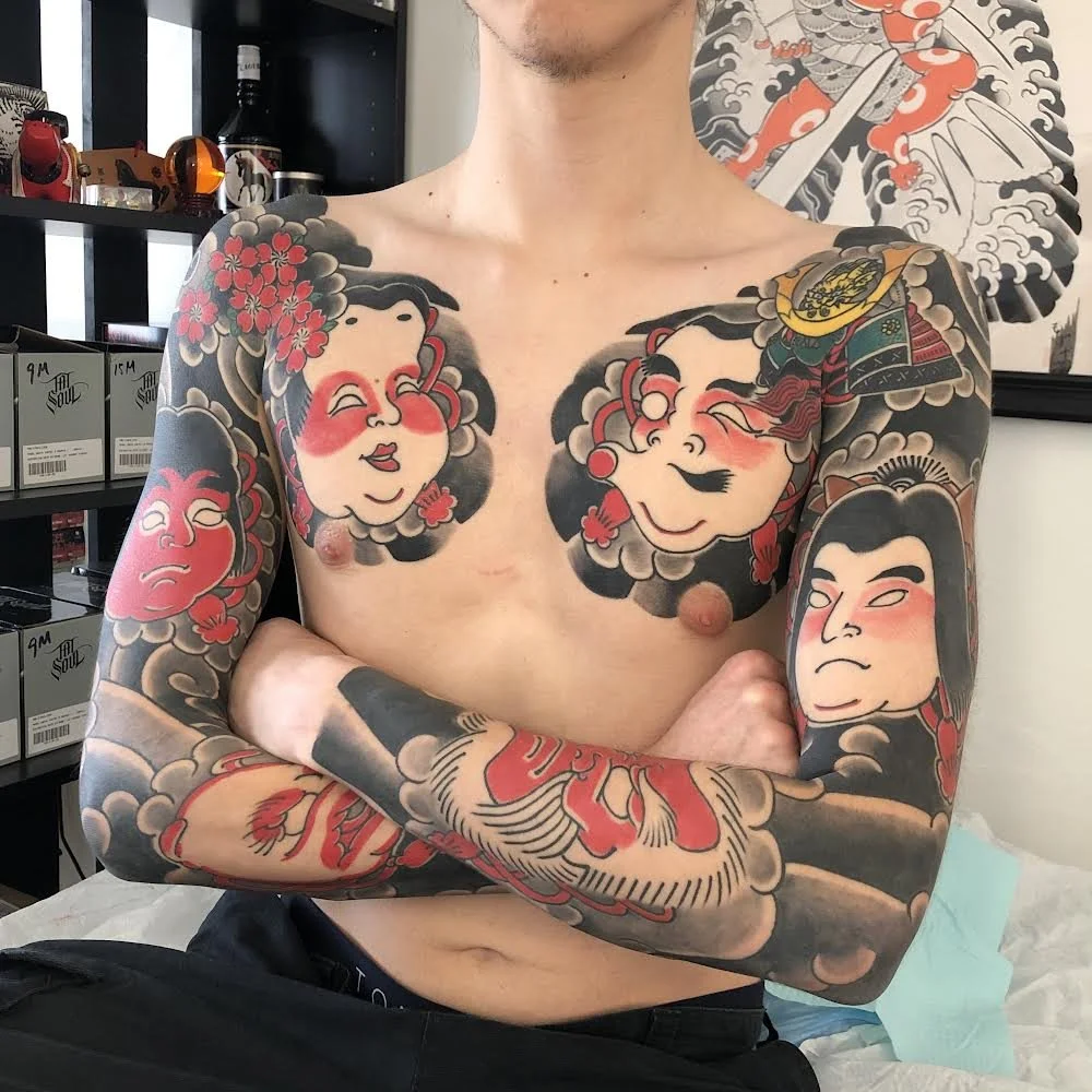 A shirtless man with extensive colorful Japanese-style tattoos on his chest and arms. The tattoos include depictions of traditional Japanese faces, a dragon, and clouds, with vivid reds, blacks, and grays.
