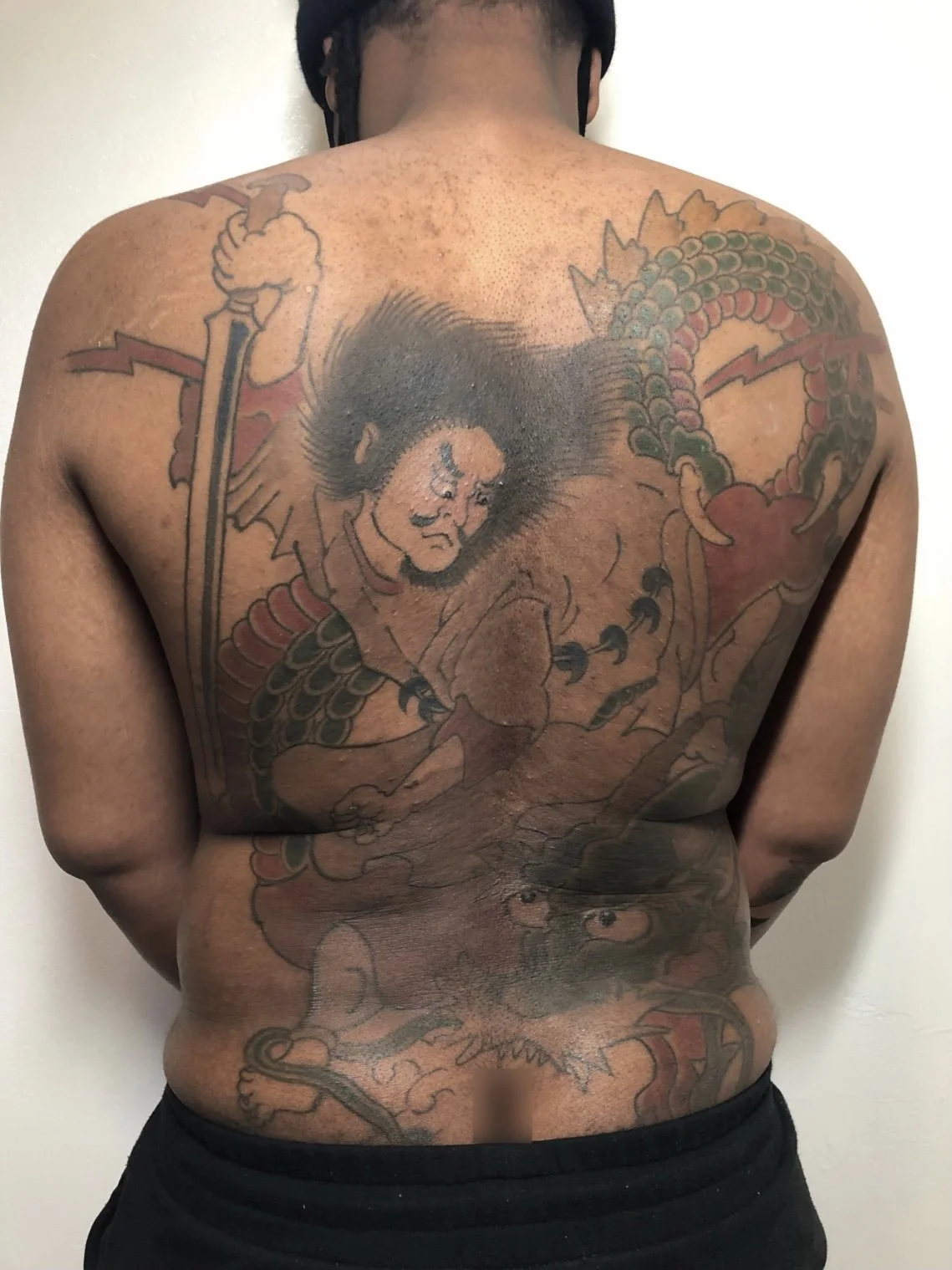 A person's back with a large colorful tattoo of a traditional Japanese-style dragon and a fierce-looking male warrior with long hair, holding a weapon, with the dragon coiling around him.