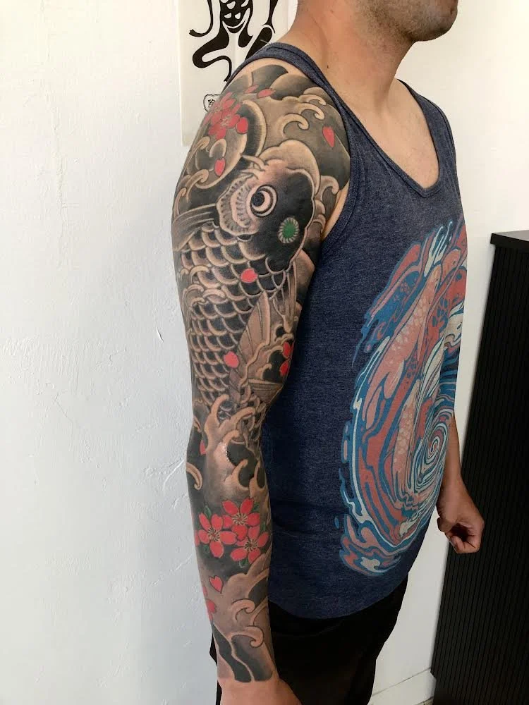 A man with a full sleeve tattoo on his left arm, featuring a large koi fish, cherry blossoms, and stylized waves, standing in a room with a white wall and a black cabinet.