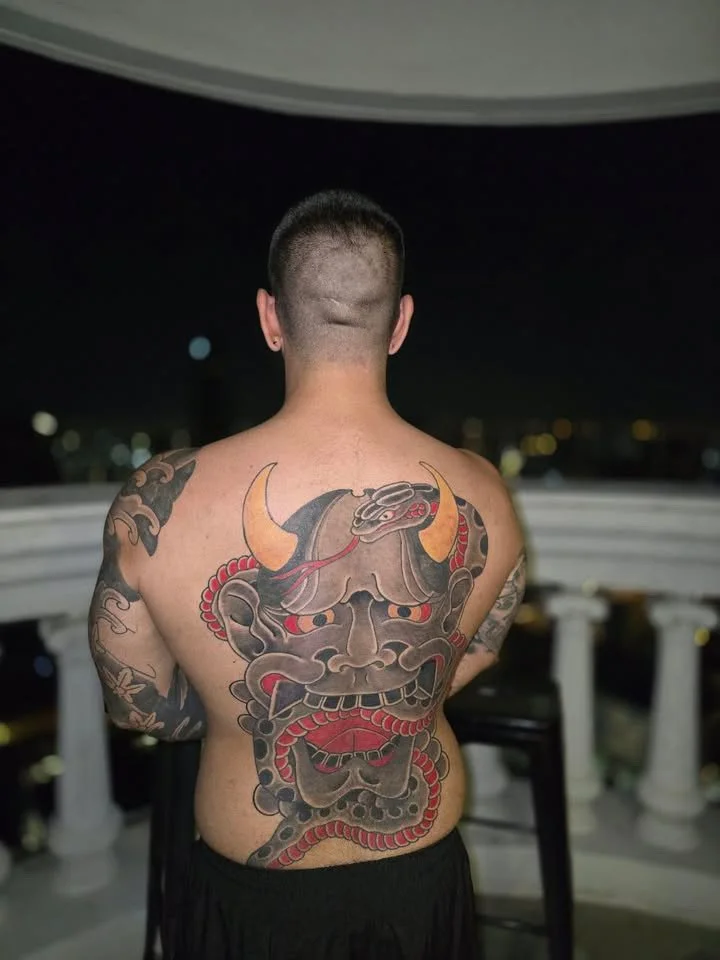 Front view of a man with a large traditional Japanese oni mask tattoo covering his back, with horns and a fierce expression.