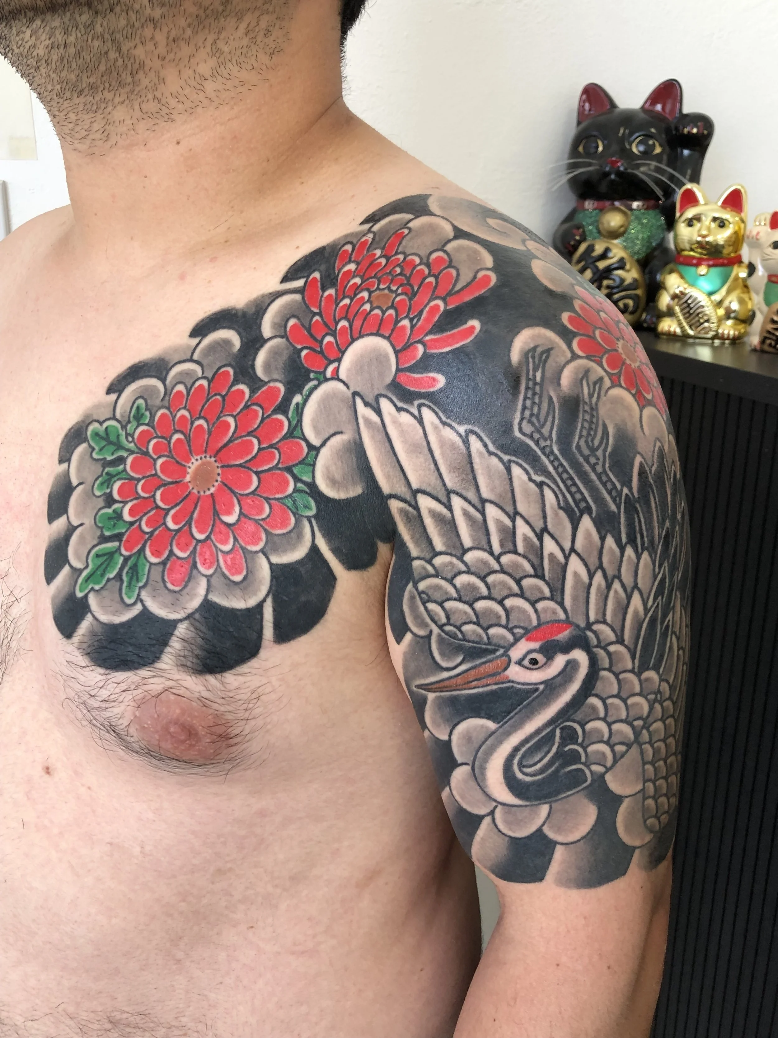 Tattoo of a heron with large red and pink flowers on the shoulder and upper arm of a person, with a background of grey clouds.