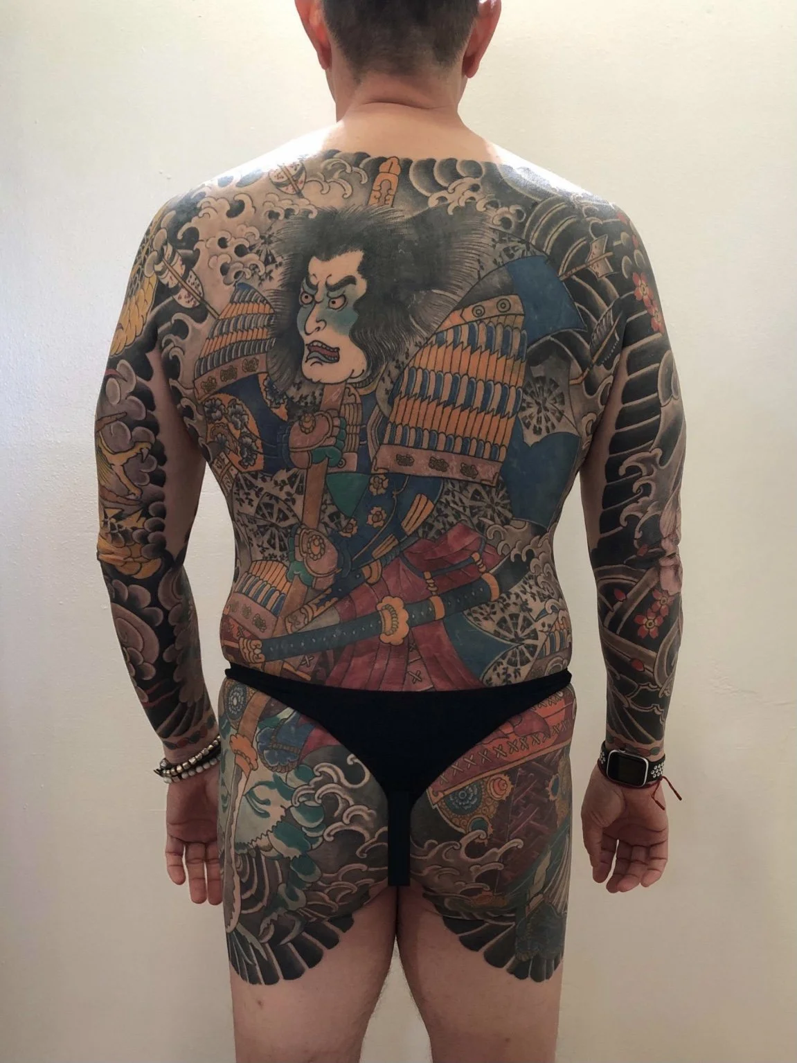 Back of a person with extensive traditional Japanese style tattoos covering the back, arms, and upper legs. The tattoo features a fierce warrior with a large face, wild hair, and armor, set amidst clouds and traditional Japanese patterns.