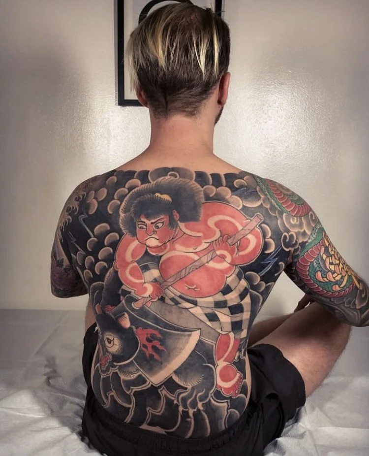 Back view of a person with detailed Japanese-style tattoo of a fierce warrior with an axe on their back, dark clouds, and a dragon on the arm, seated on a bed against a plain wall.