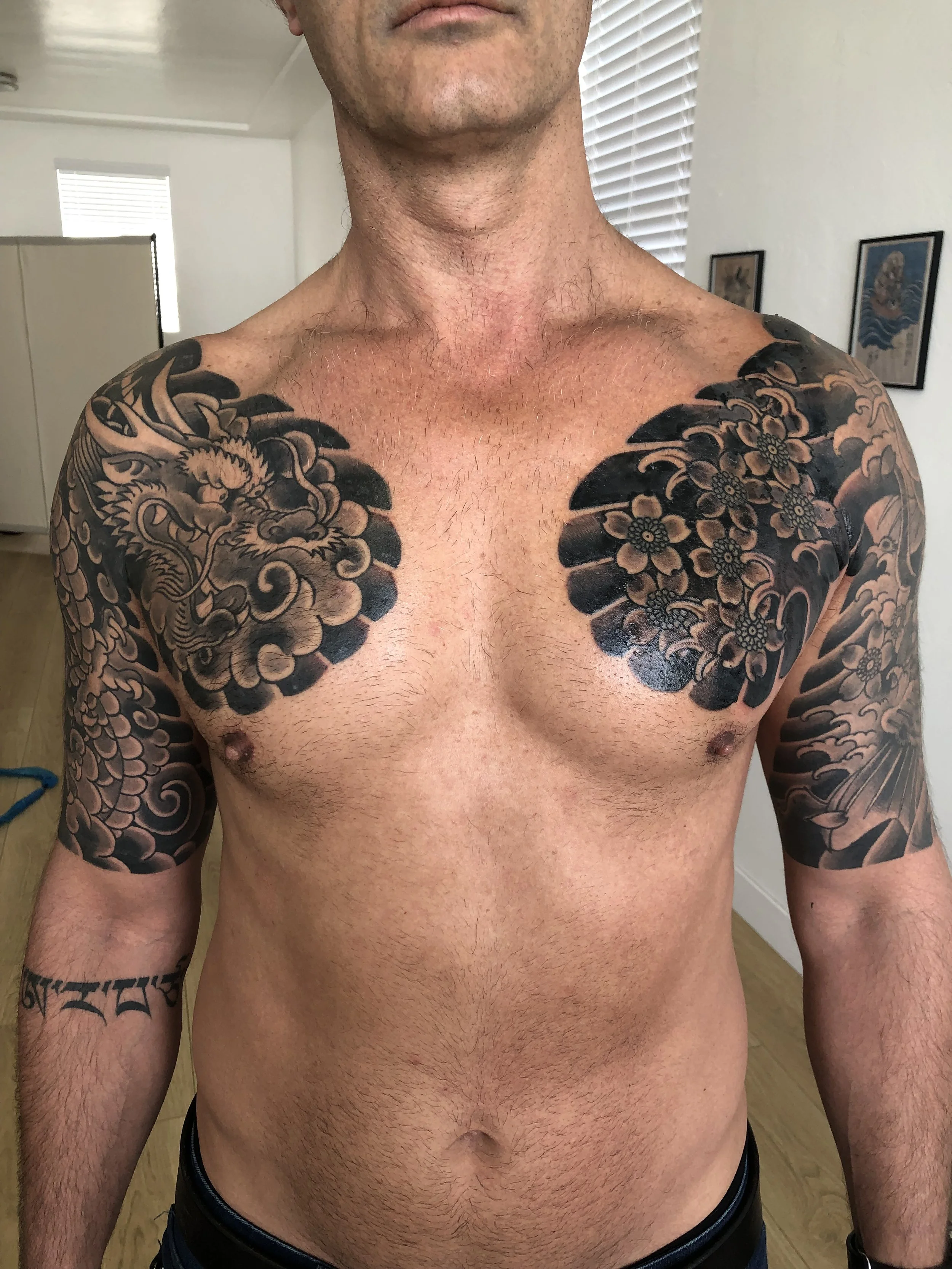 A shirtless man showing detailed black tattoo art on his chest, shoulders, and arms, with a wooden floor and framed pictures on the wall in the background.