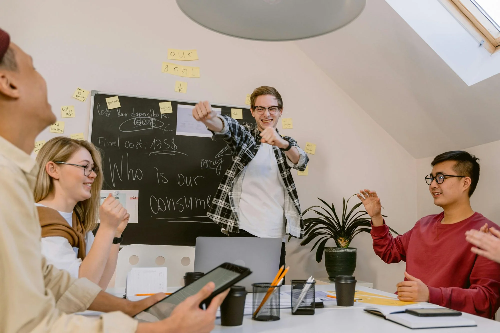 Four people in a meeting room discussing ideas as one person gestures toward a blackboard filled with handwritten notes and sticky notes.