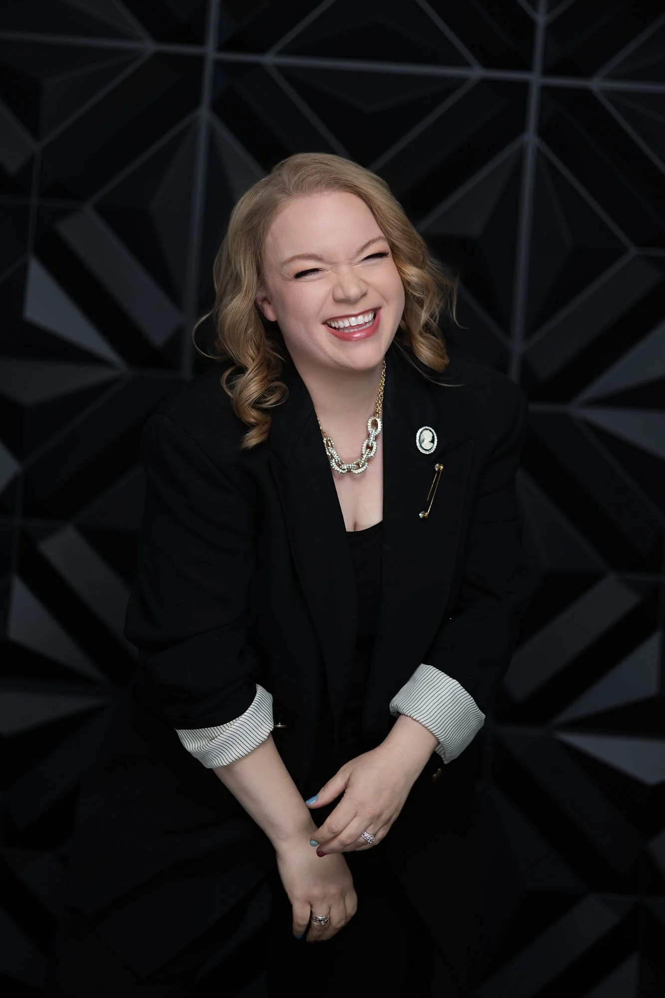 Jessie Hemmelgarn laughing for the camera in a black business suit in front of a black textured background. Photo by Desired Focus Photography