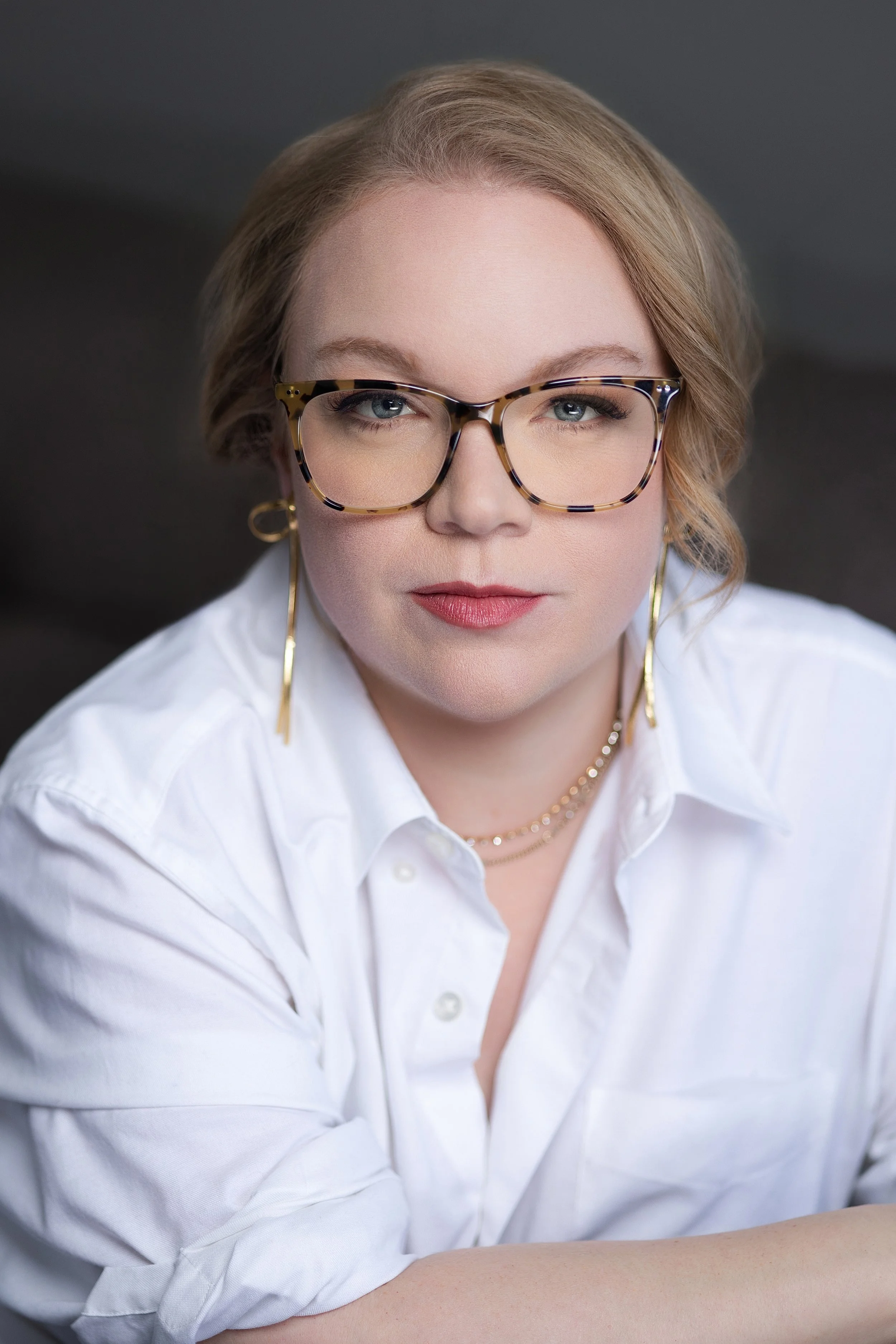 Close-up of a woman with blonde hair, wearing tortoiseshell glasses, gold earrings, and layered necklaces, dressed in a white shirt against a dark background.