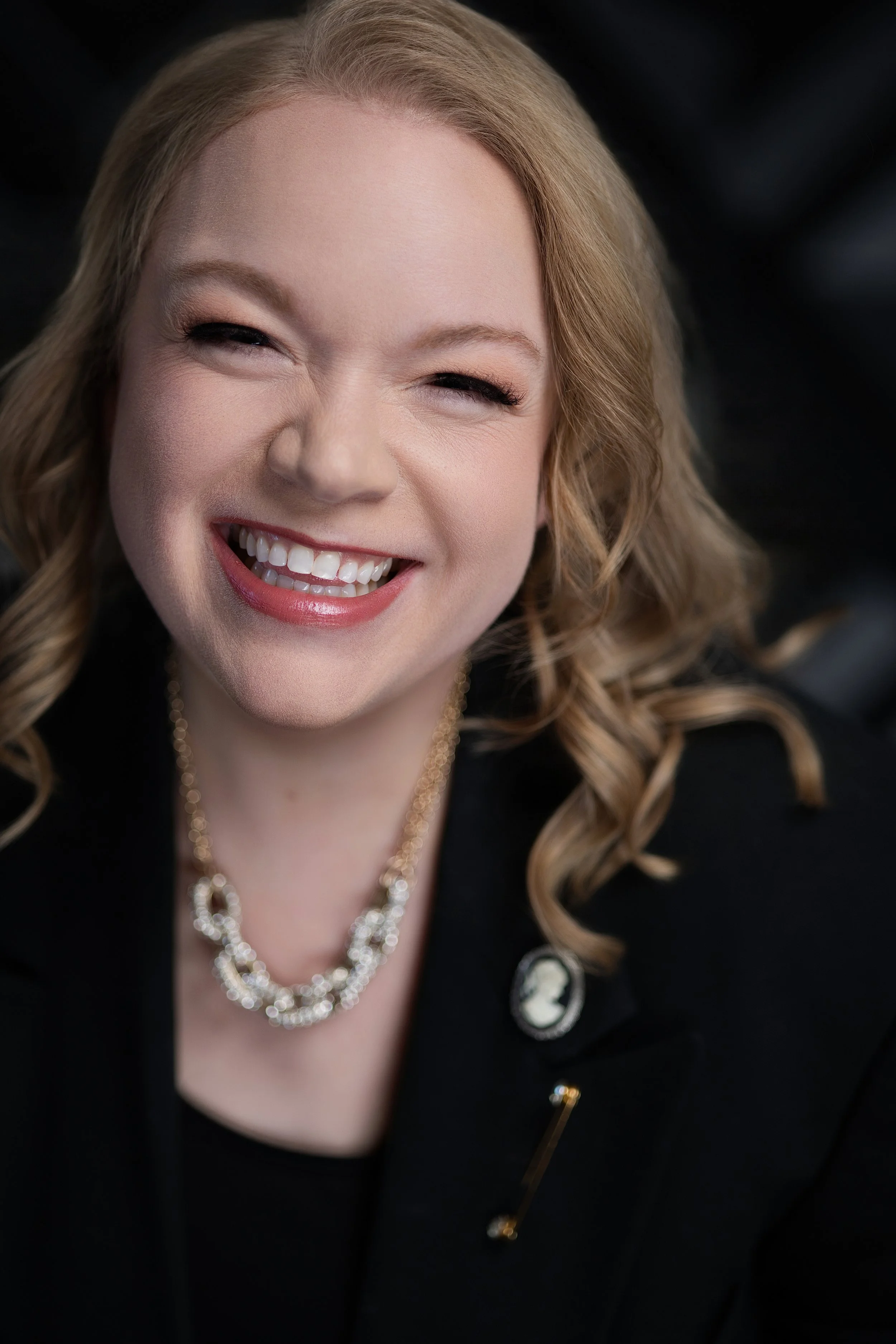 Close-up of Jessie Hemmelgarn of Wonderline Creative wearing a black blazer, a pearl necklace, and a brooch on her lapel, against a dark background.