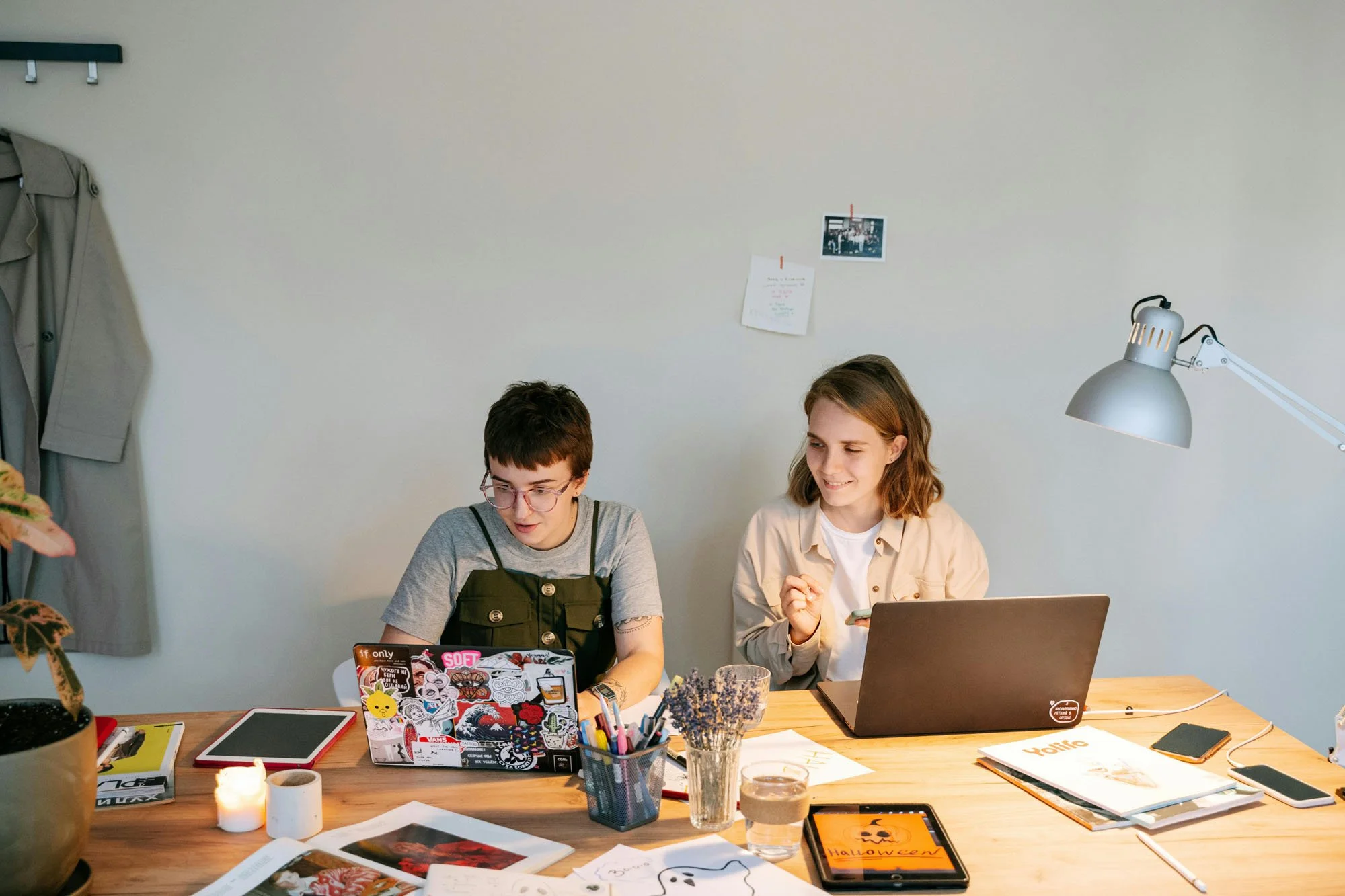 Two people sitting at a wooden desk with laptops, notebooks, printed images, a tablet illustration, and various creative supplies, working together in a warmly lit workspace.