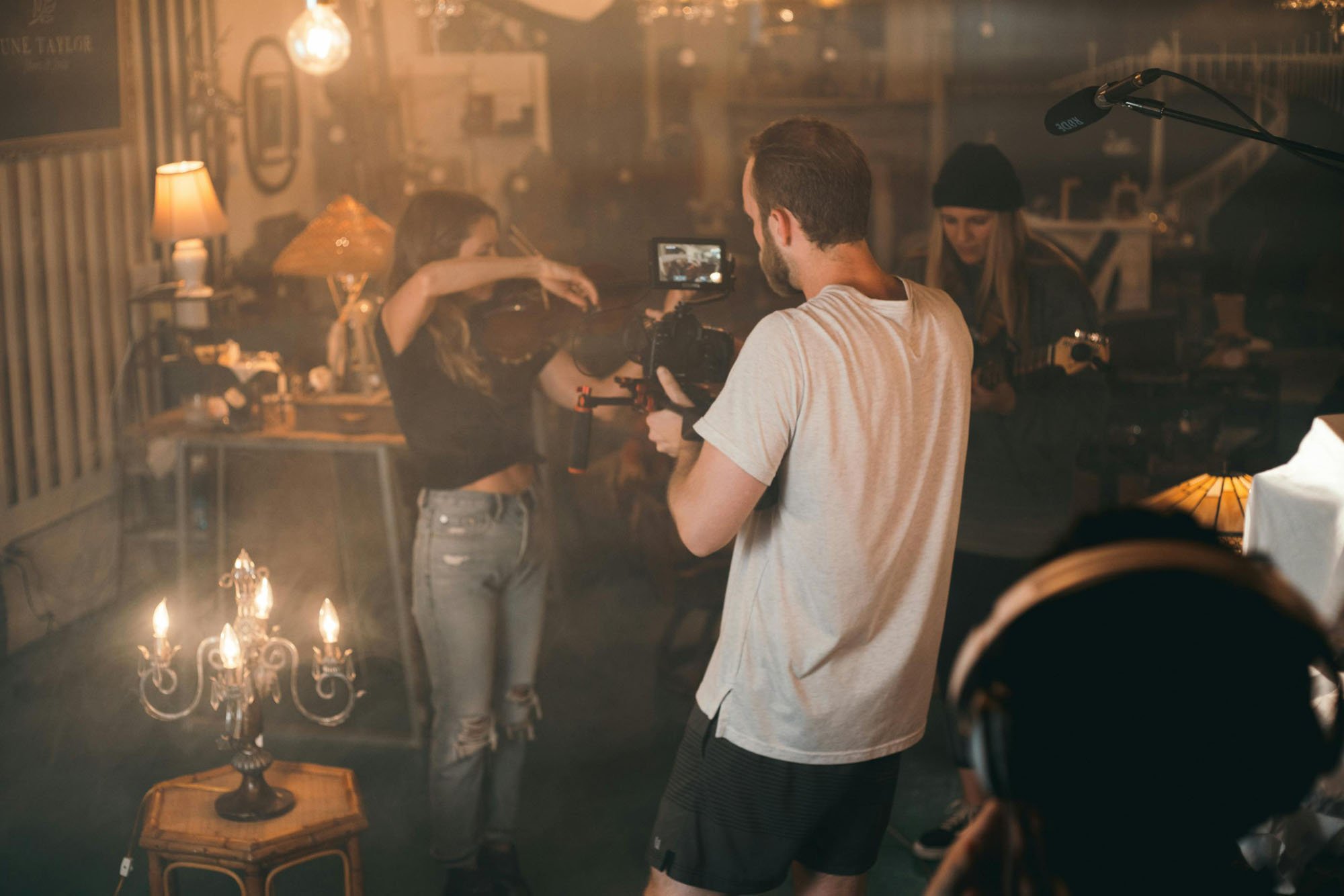 A videographer operates a professional camera while filming a violinist and another musician in a warmly lit studio filled with lamps, props, and audio equipment.