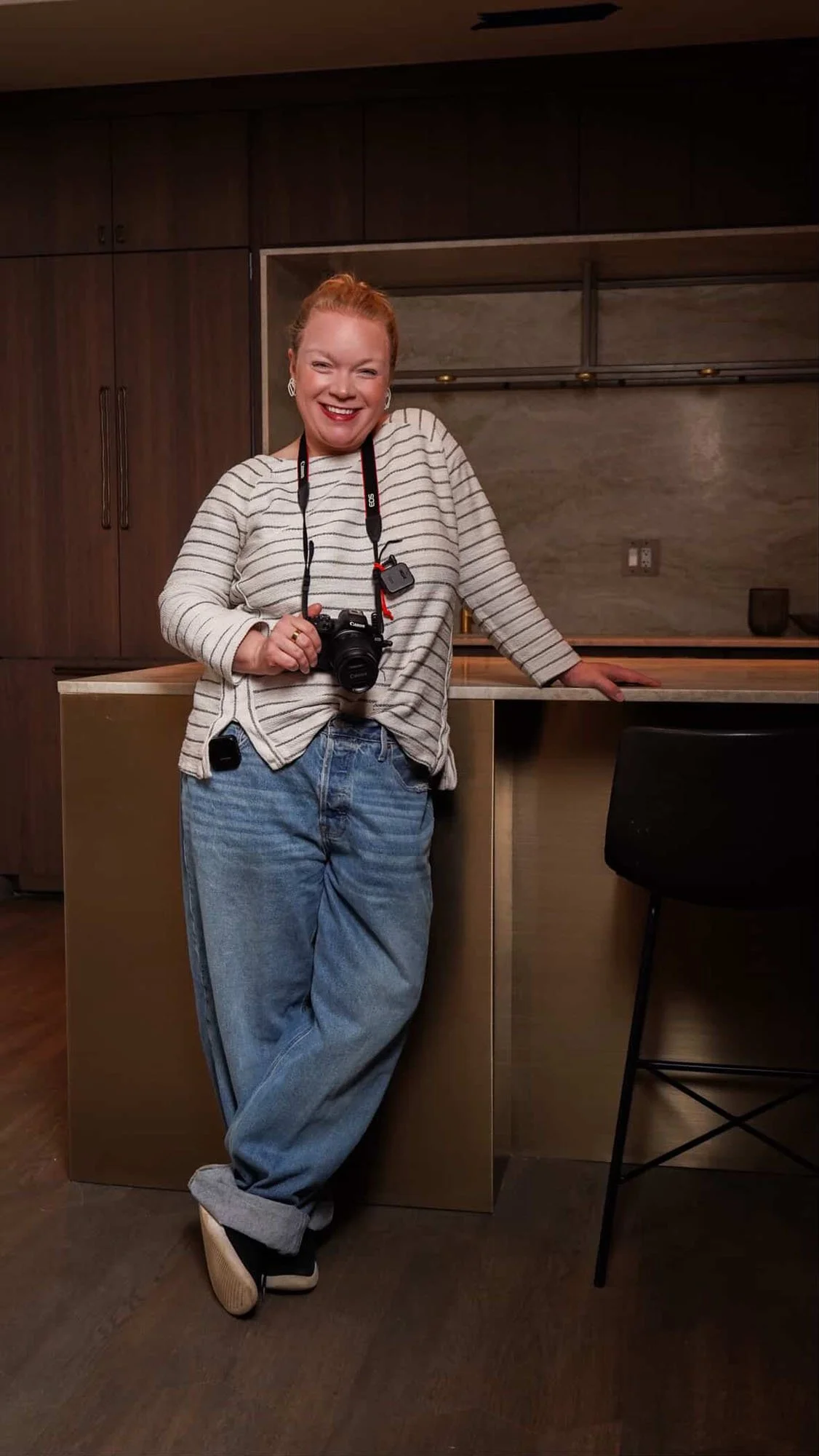Jessie Hemmelgarn wearing a striped shirt and loose jeans, stands in a modern kitchen, smiling while holding a camera around her neck.