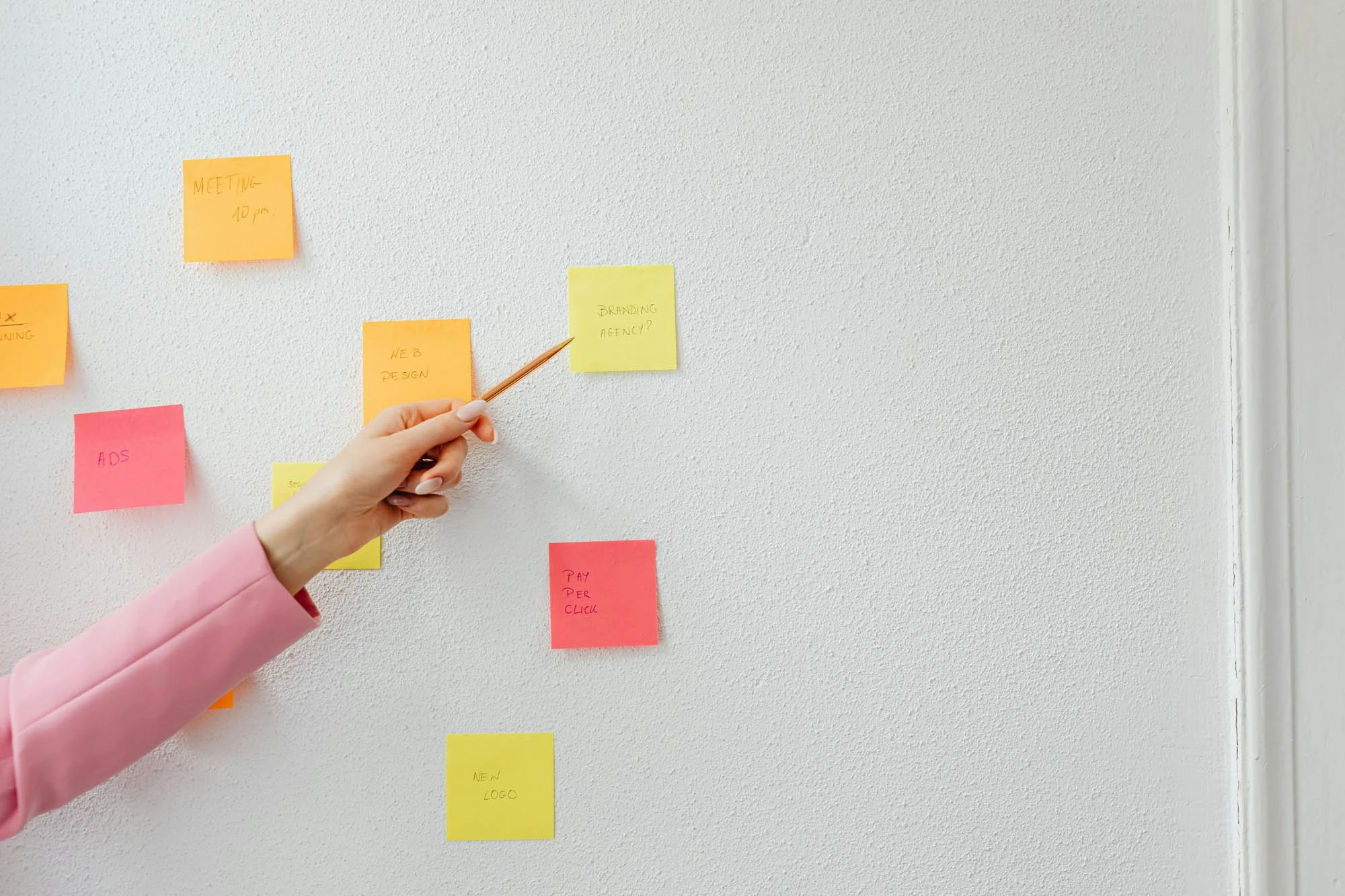 A hand in a pink sleeve points to colorful sticky notes on a white wall, each labeled with tasks and ideas for a creative project.