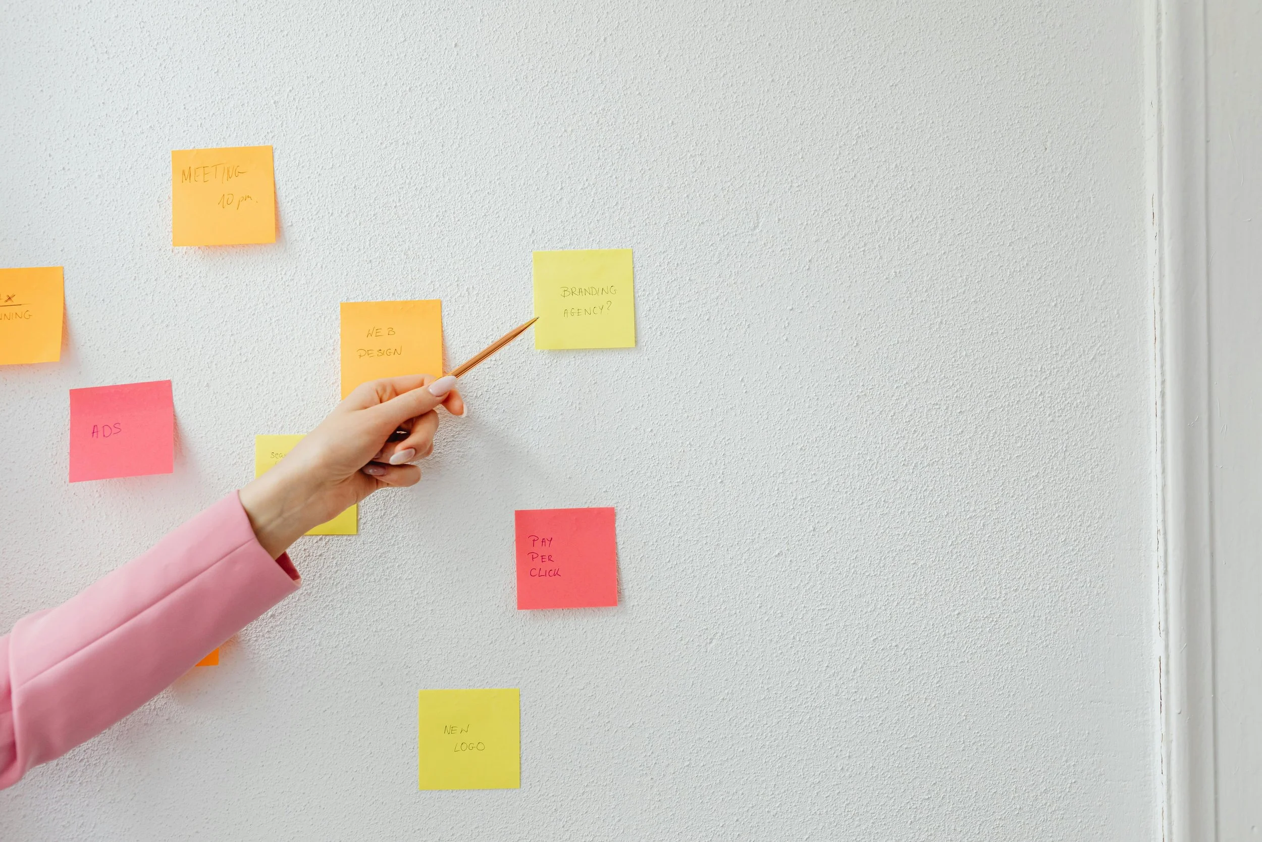 Person in pink blazer attaching a yellow sticky note labeled 'BRANDING AGENCY?' to a wall with several other colorful sticky notes.