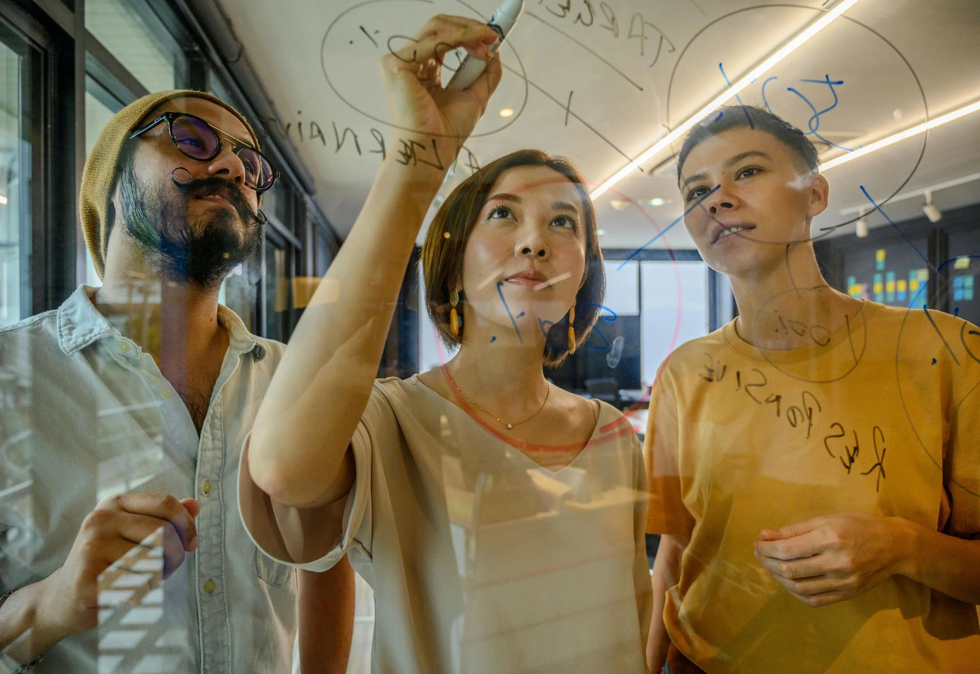 Three team members brainstorming at a glass board, sketching concepts and aligning on next‑step strategy.