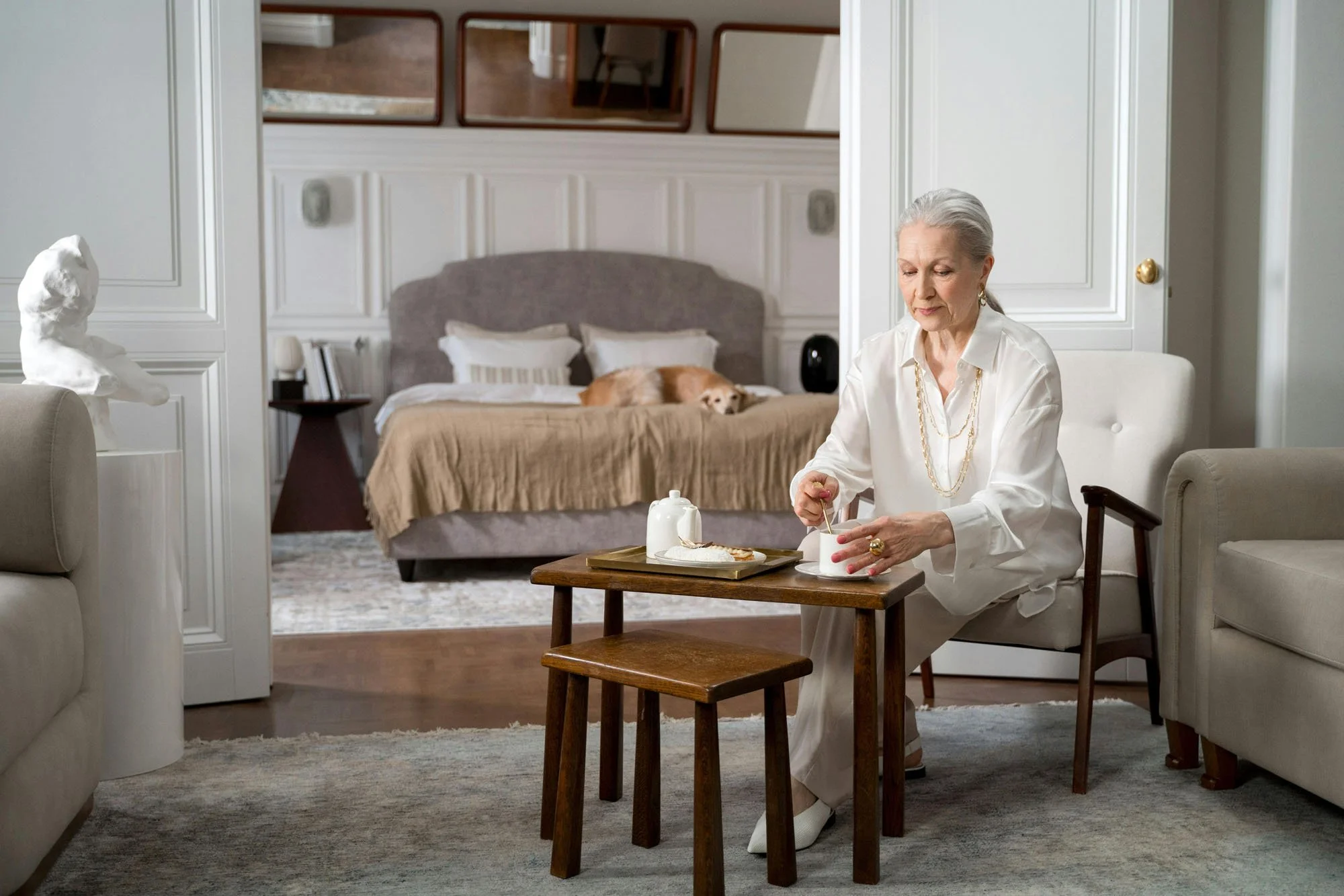 An older adult relaxing in an elegant bedroom sitting area, stirring a drink beside a tray with tea and a pastry.