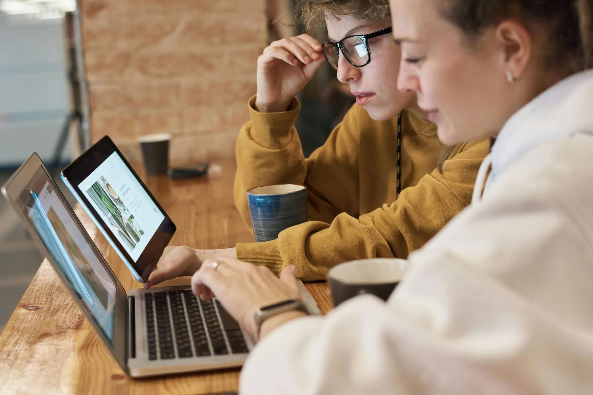 Two people sitting at a wooden table with laptops and coffee mugs, concentrating on their work in a casual workspace with a brick wall in the background.