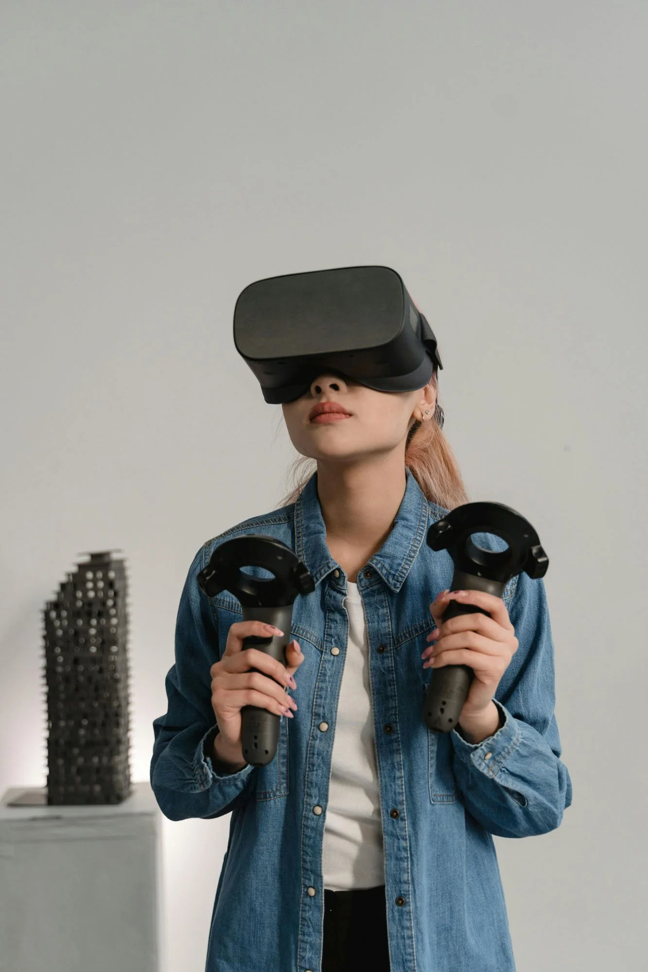 A person wearing a black VR headset and holding two VR controllers stands in front of a light background, with a sculptural model displayed on a pedestal behind them.