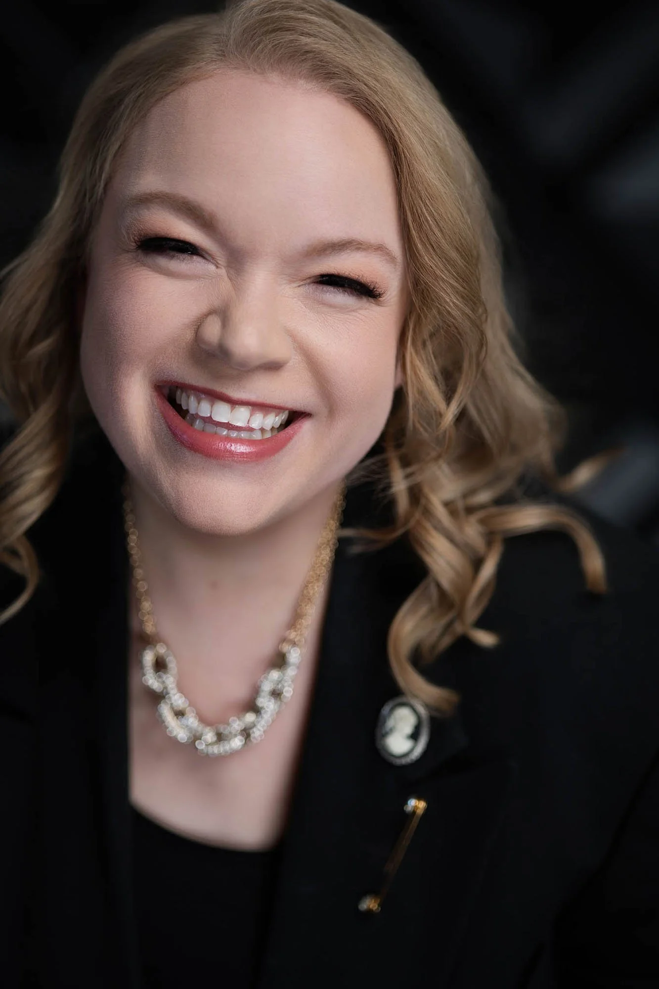 Close-up of Jessie Hemmelgarn of Wonderline Creative wearing a black blazer, a pearl necklace, and a brooch on her lapel, against a dark background.