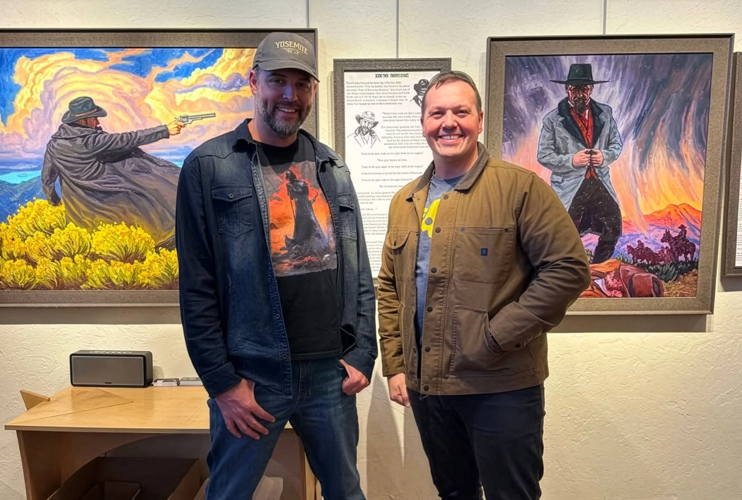 Two men standing in art gallery in front of colorful paintings depicting cowboys, with a side table and speaker nearby.