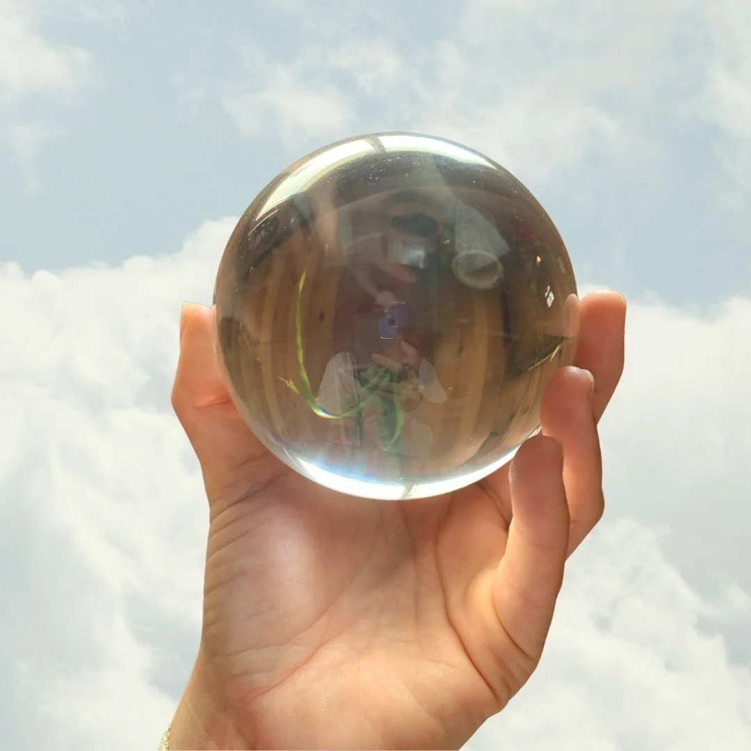 A hand holding a clear glass sphere against a cloudy sky, with a reflection of the person taking the photo visible inside the sphere.