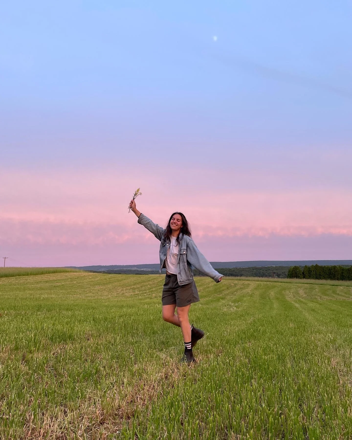 A young woman in casual clothes standing in a grassy field during sunset, holding a small bouquet of flowers with her right hand raised, smiling happily.