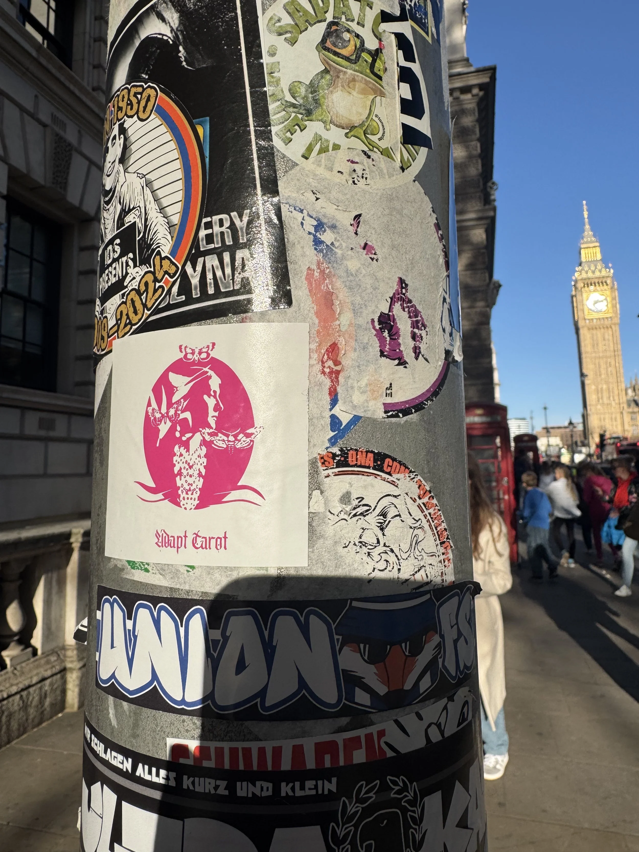 A pole covered in various stickers with Big Ben clock tower and people walking on the street in the background.