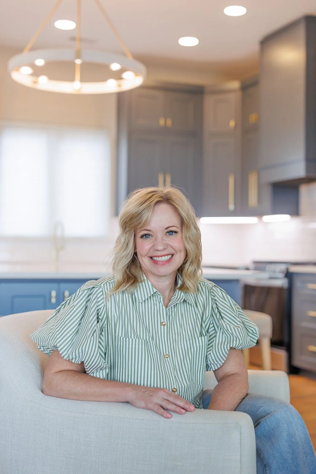 A smiling woman with blonde hair in a green and white striped shirt sitting on a light-colored couch in a modern kitchen with blue cabinets and gold hardware.