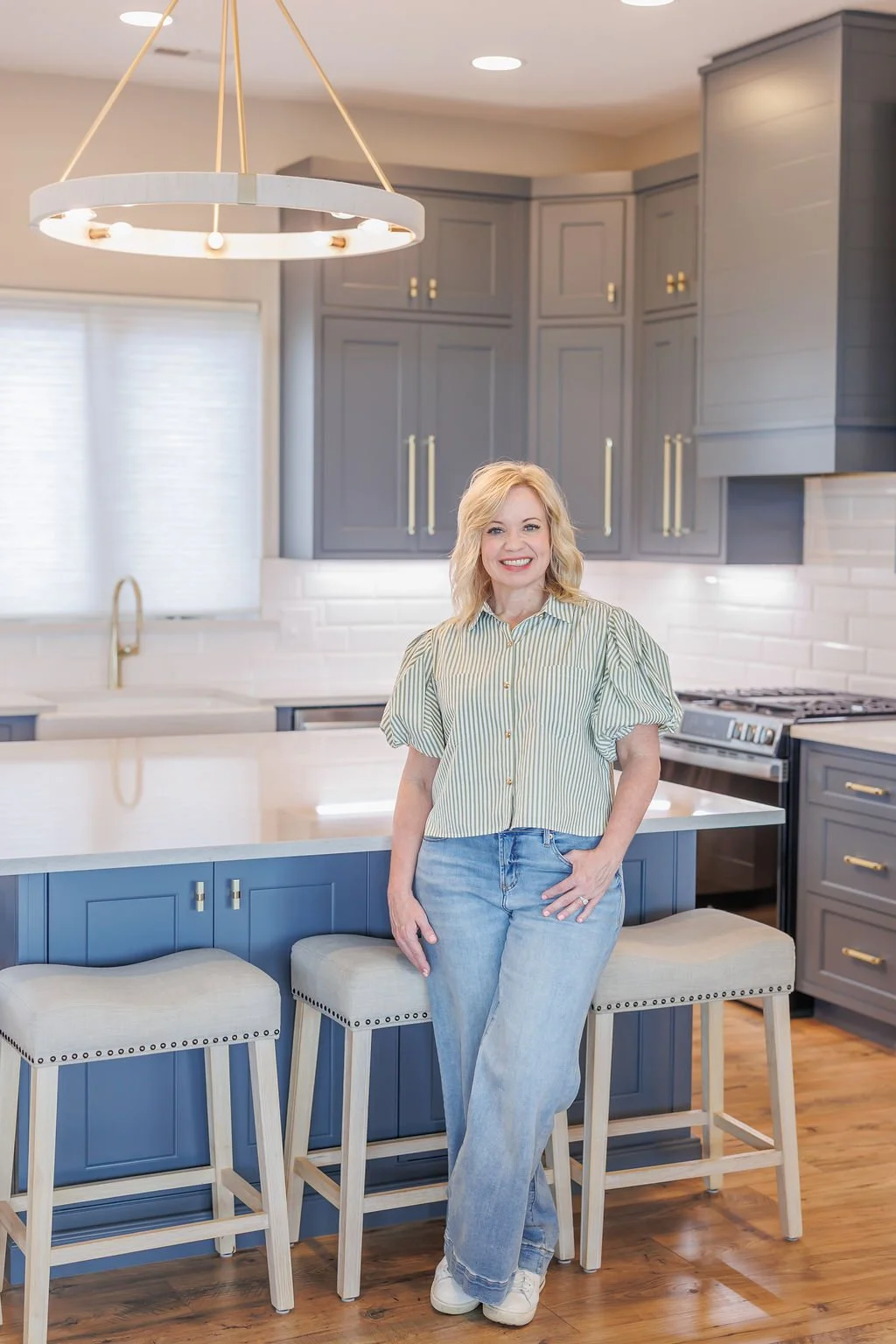 A woman with blonde hair smiling and standing in a modern kitchen with gray cabinets, white countertops, and a large island. She is wearing a striped shirt and jeans, with kitchen stools in front of the island.