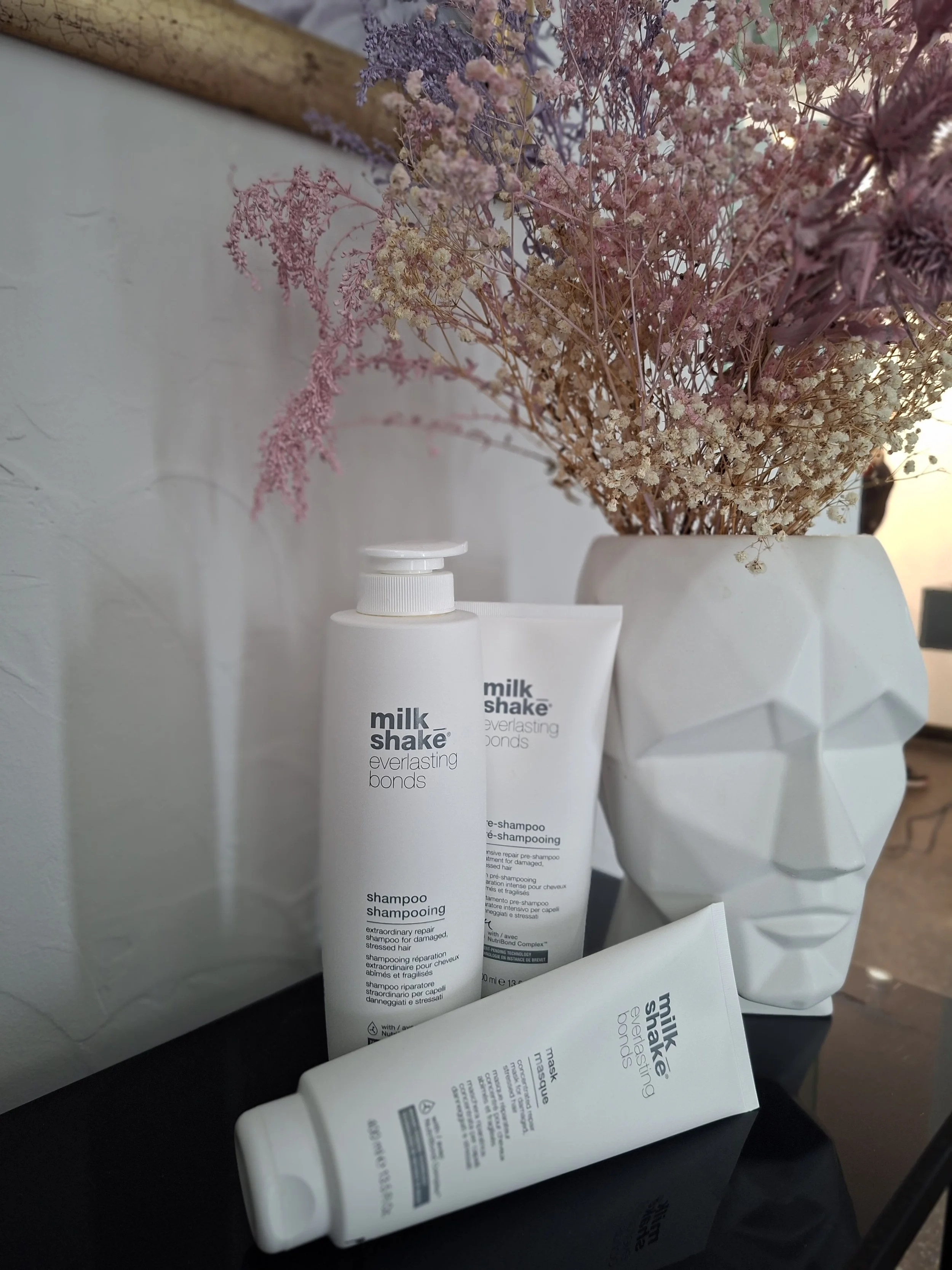 A white bottle of shampoo labeled 'milk shake everlasting bonds' sitting on a black surface, with dried pink, purple, and beige flowers in the background in a white vase.