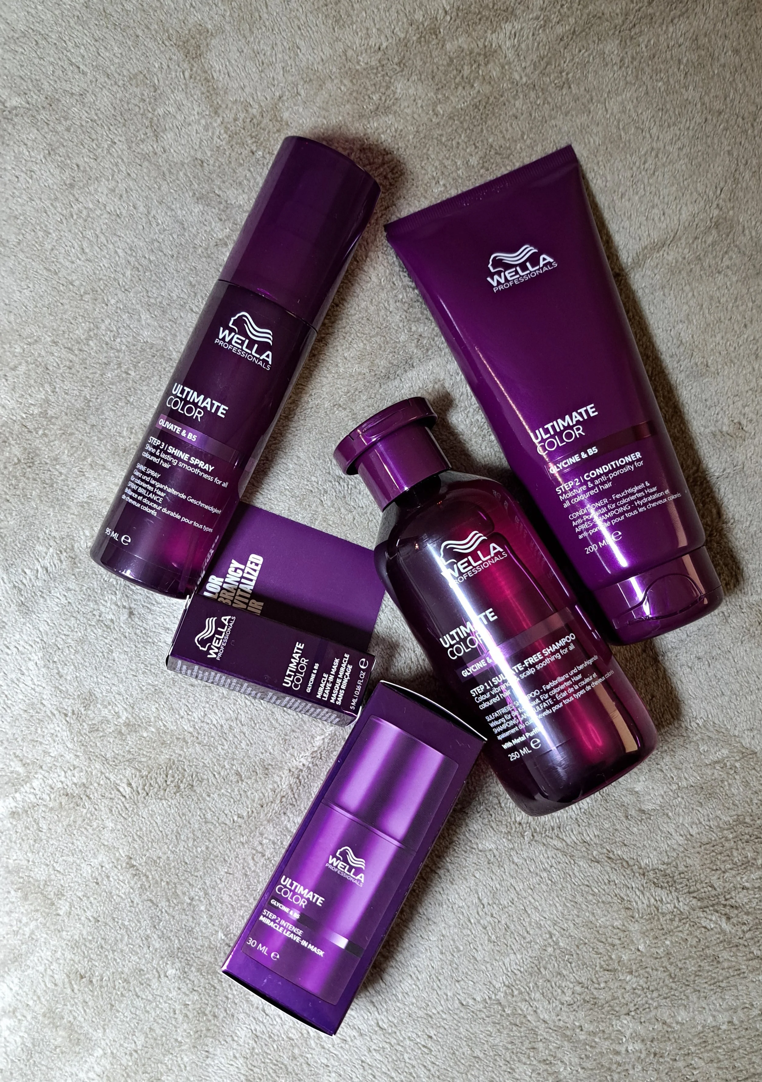 Collection of Wella Professionals Ultimate Color hair care products, including shampoo, conditioner, leave-in mask, and spray, all in purple packaging on a beige carpet.