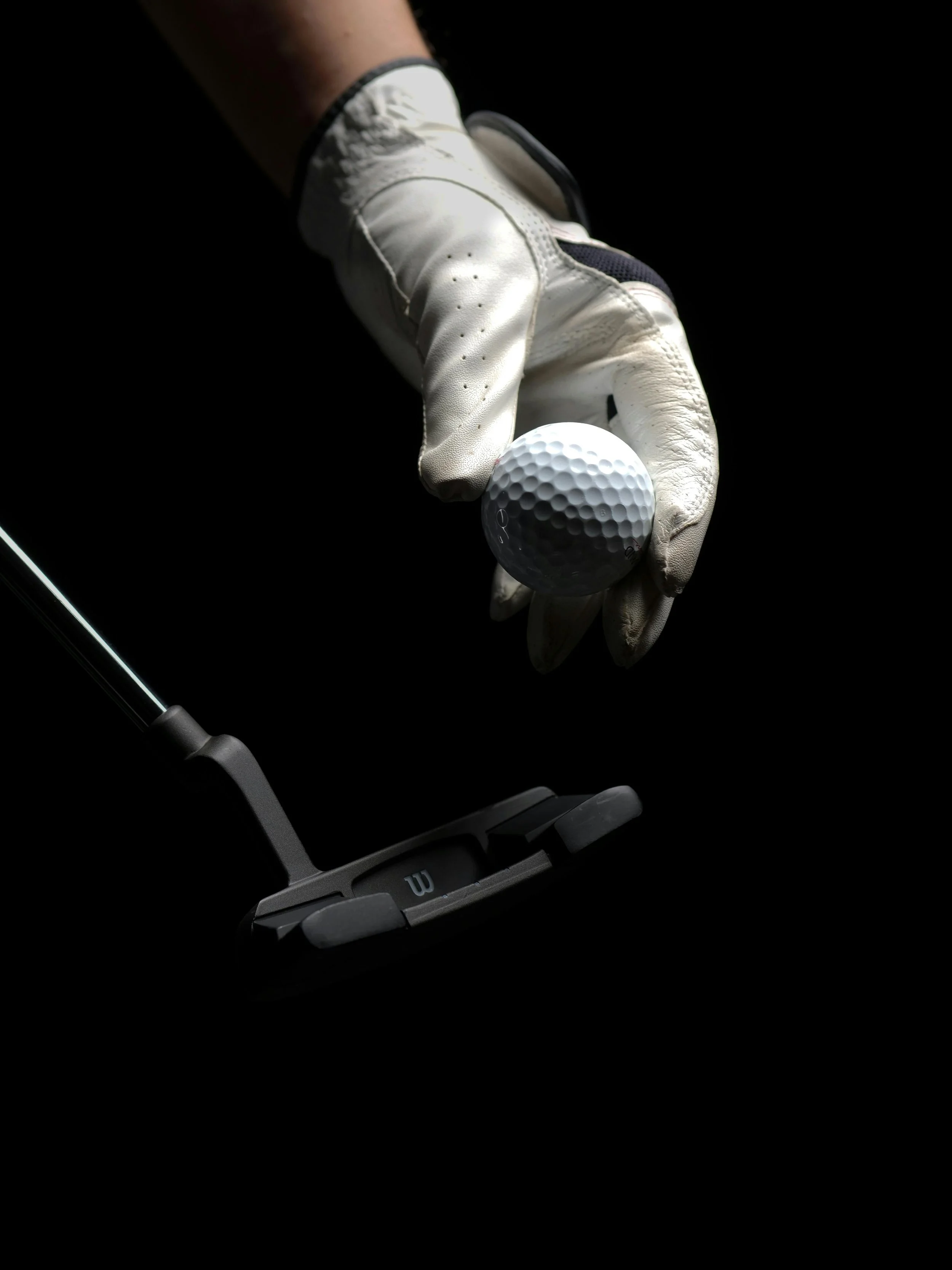 A golfer wearing a white golf glove holding a white golf ball, with a putter below against a black background.