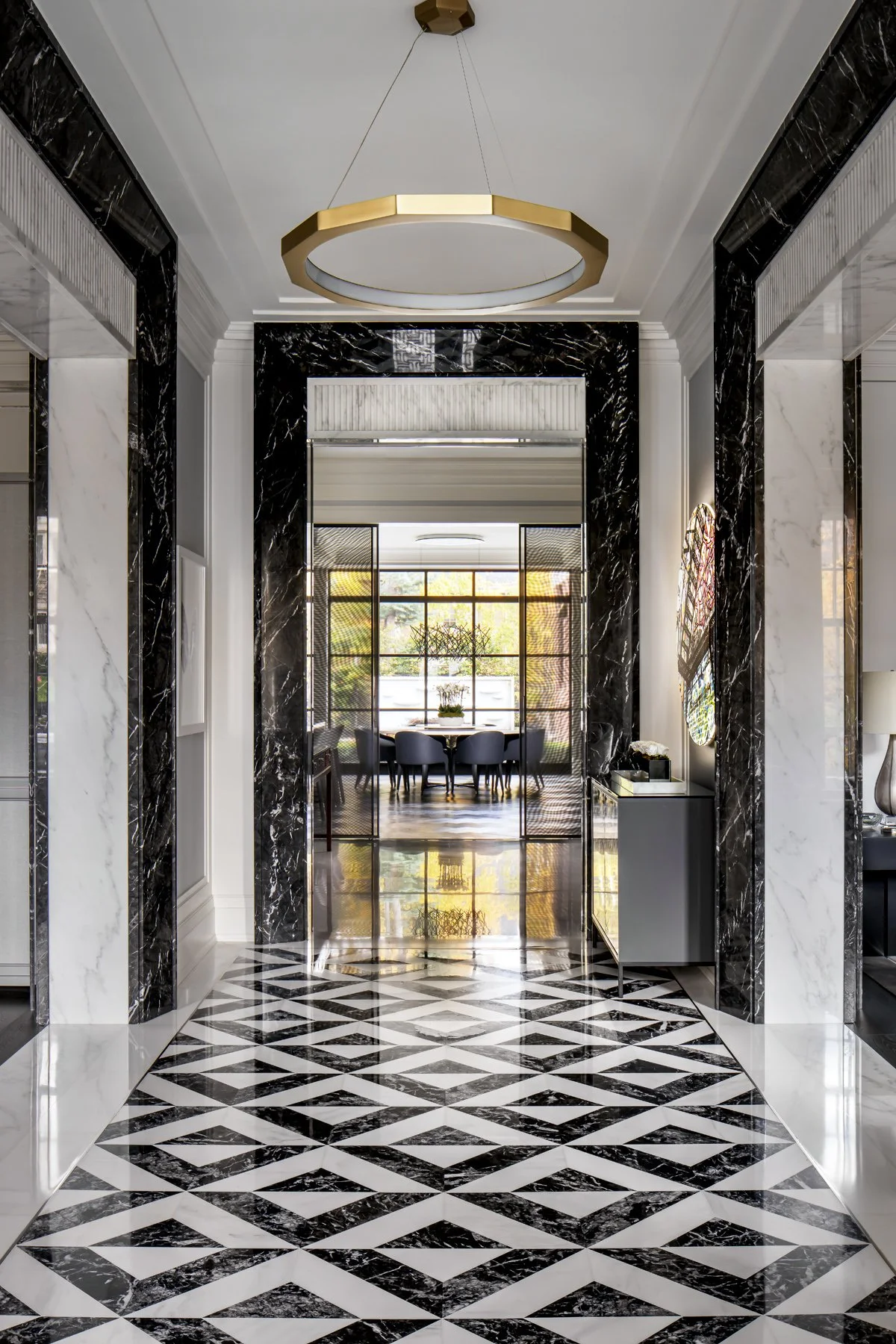 Elegant hallway with black and white marble flooring and black marble wall accents leading to a dining room with large windows and a round table surrounded by gray chairs.