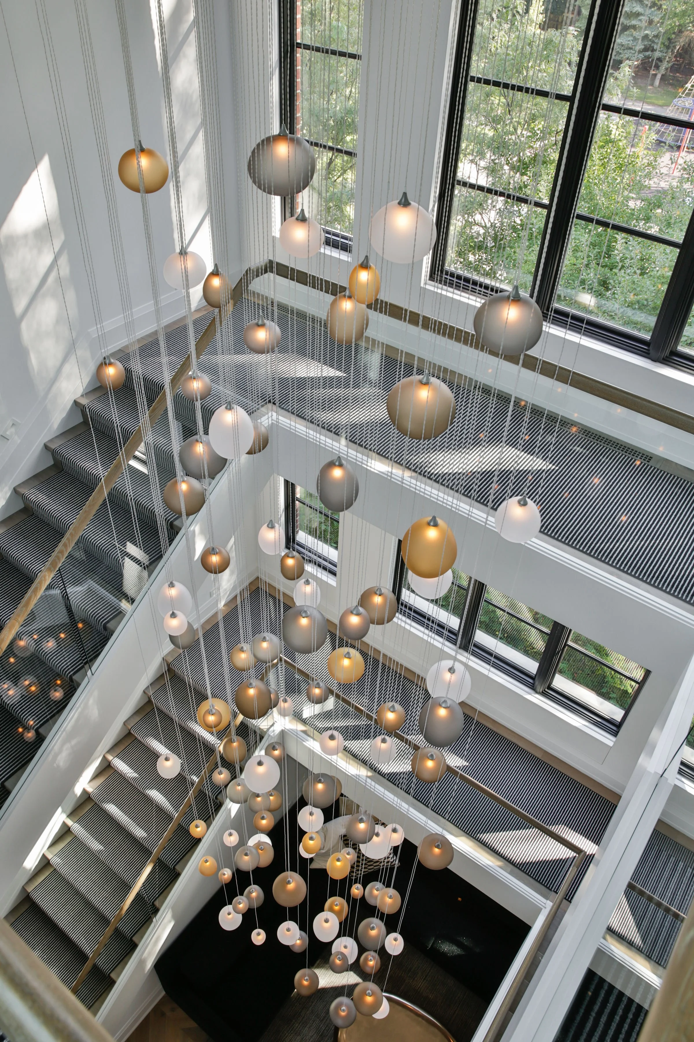 A view looking down at a modern, multi-story staircase with hanging pendant lights and large windows showing trees outside.