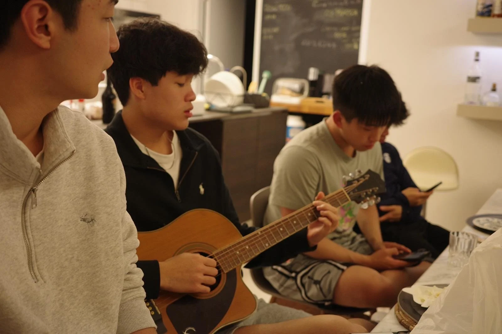 A group of young men sitting at a table, with one playing an acoustic guitar and others using their phones in a cozy indoor setting, possibly a kitchen or dining area.