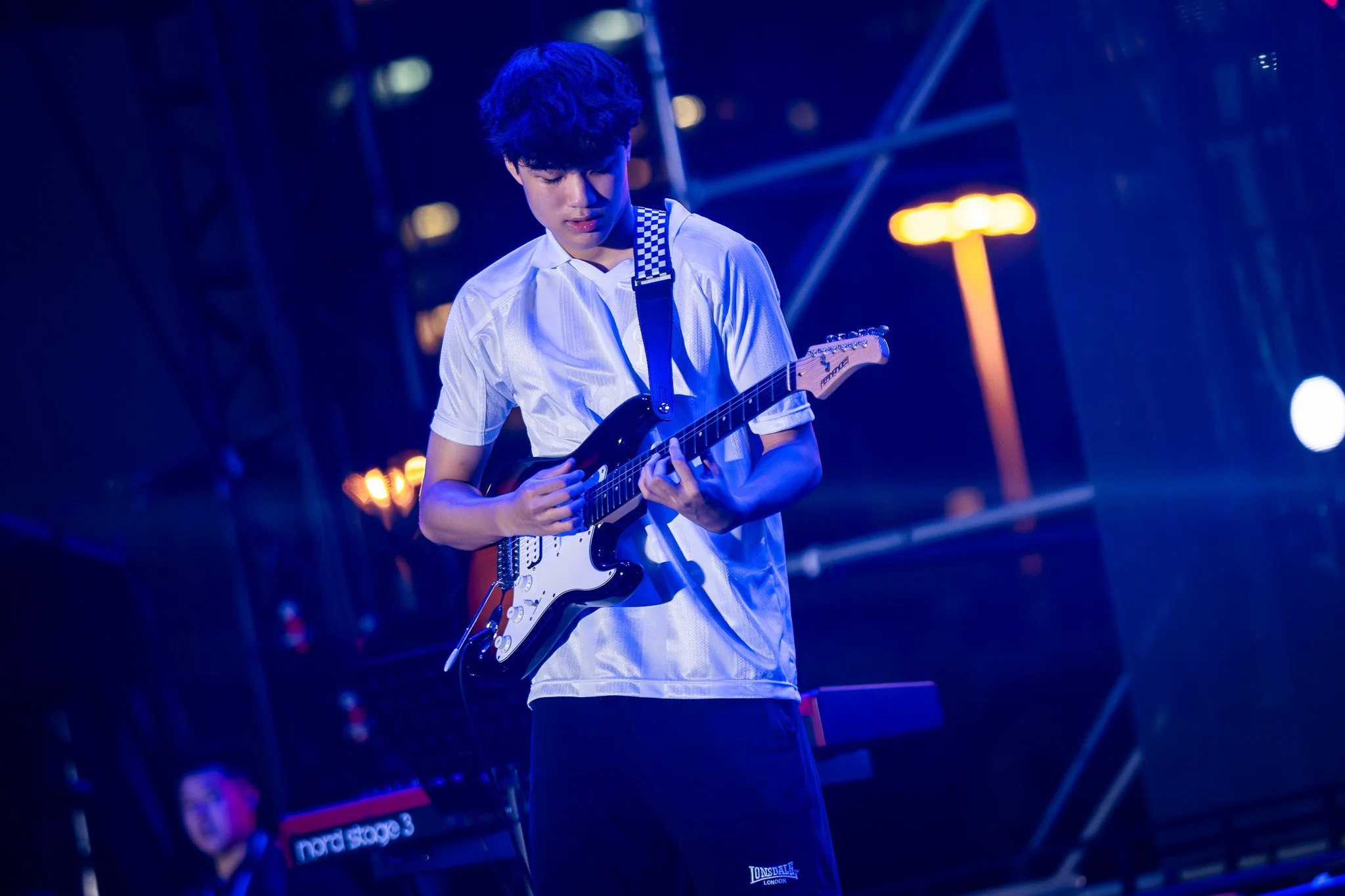 A young man with dark curly hair playing an electric guitar on stage under blue and orange lighting.