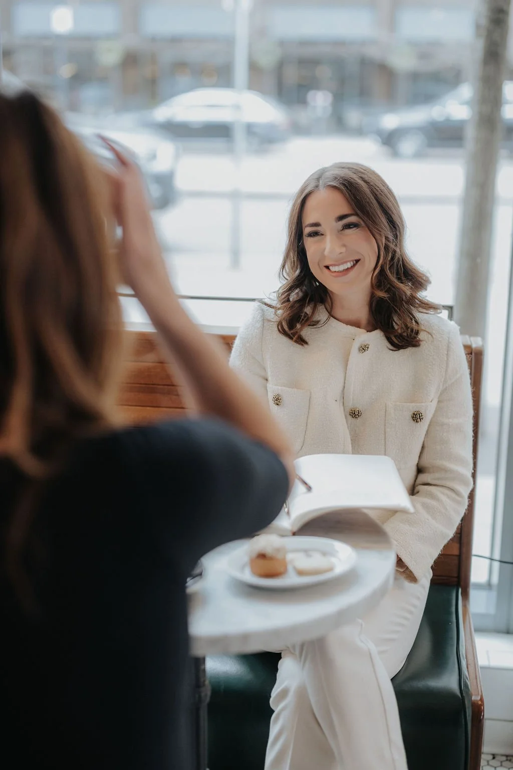 Two women having a conversation at a cafe, one smiling and holding a book, with a plate of cookies on the table.