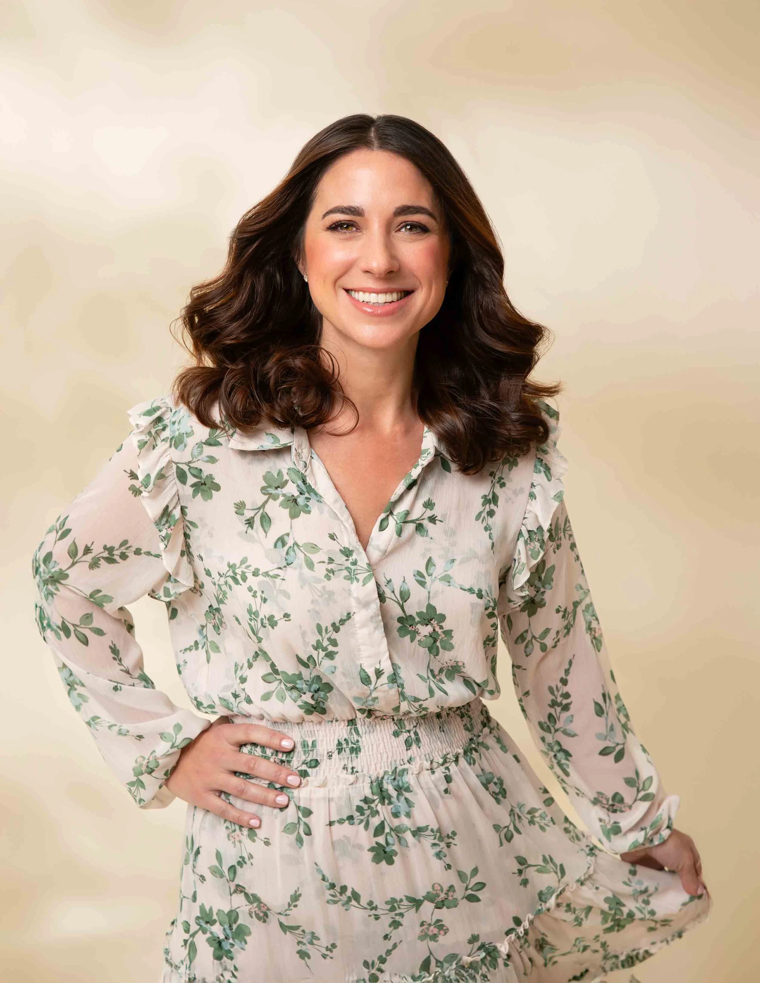 A woman with wavy brown hair wearing a cream-colored dress with green leaf patterns, smiling and posing with her hand on her hip against a beige background.
