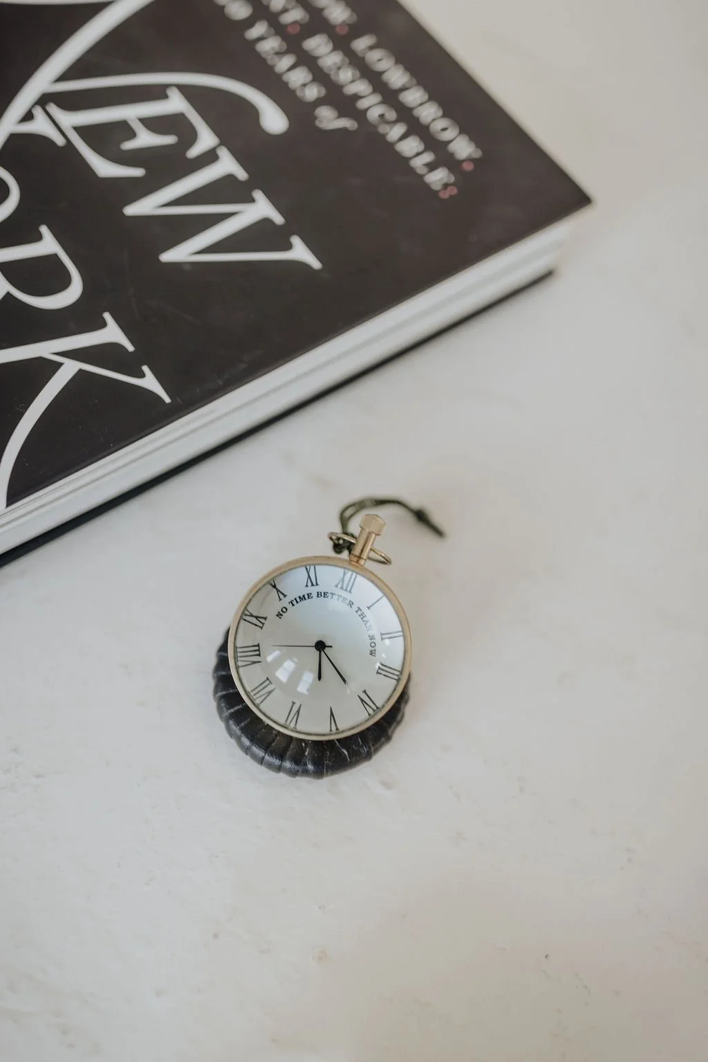 A pocket watch with Roman numerals and the phrase "NO TIME BETTER THAN NOW" on its face, lying on a white surface near a book.