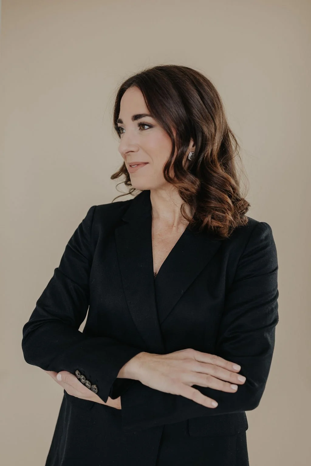 A woman with shoulder-length brown hair styled in loose waves, wearing a black blazer with buttoned sleeves, standing against a beige background. She is looking to her left with a slight smile and has her arms crossed.