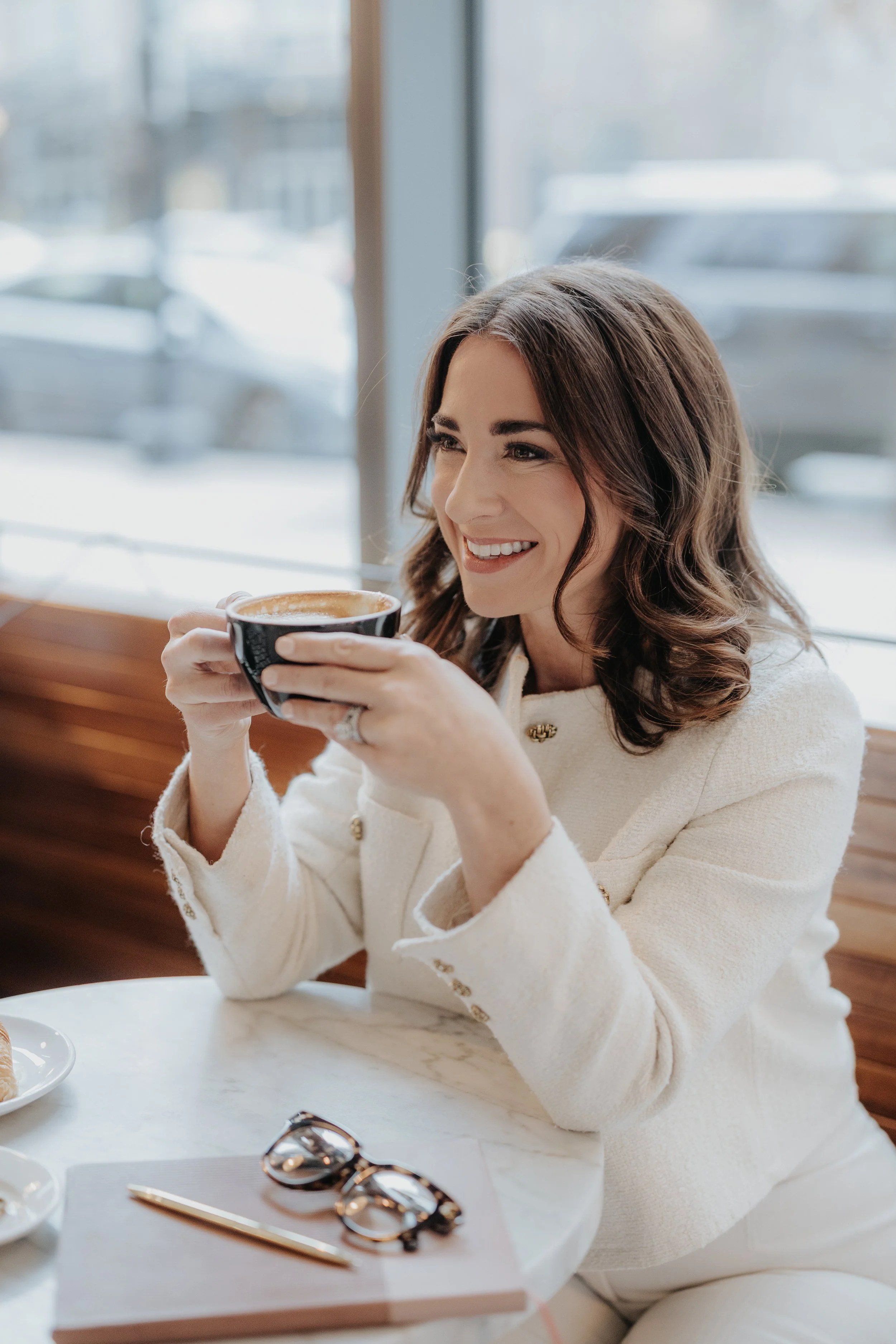 A woman with brown hair smiling while drinking coffee in a cozy cafe with large windows and a city view.
