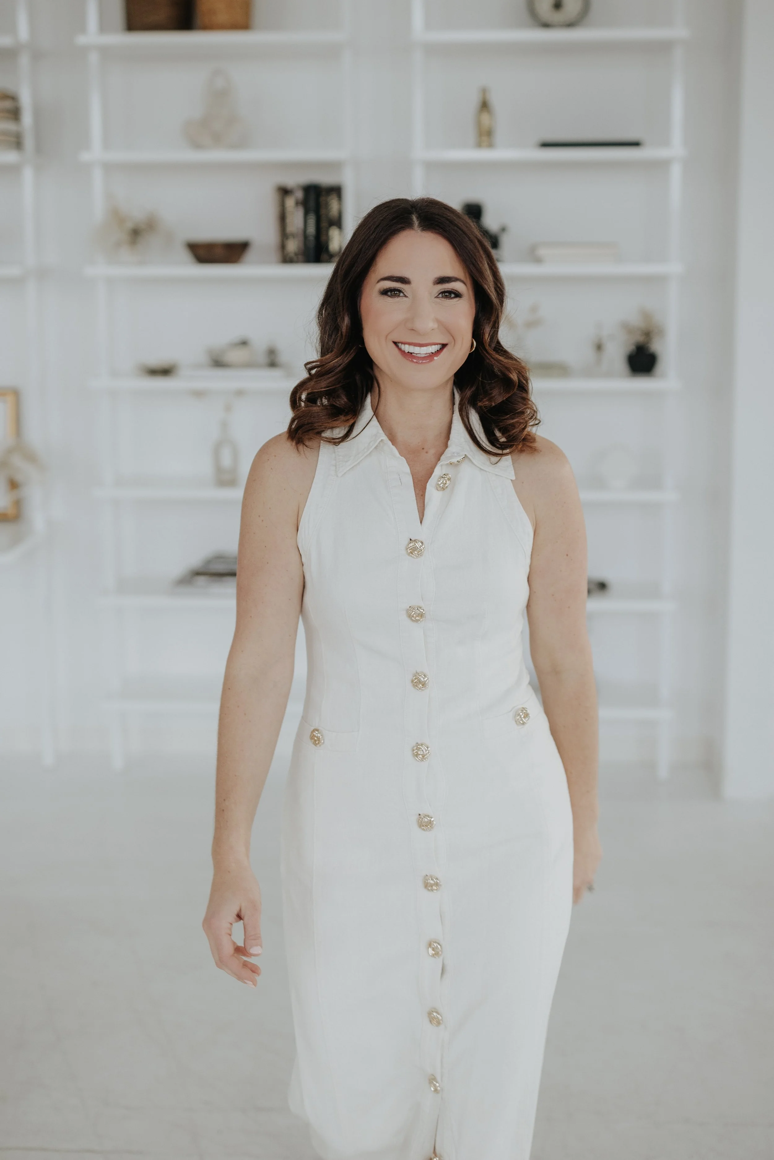 A woman smiling in front of white shelves with decorative items in a bright room.