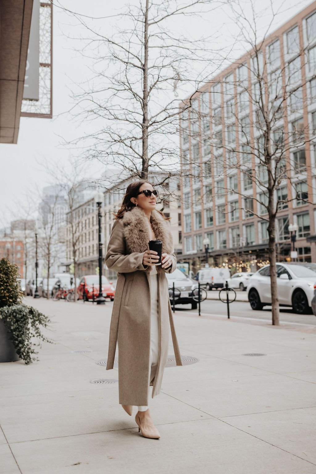 A woman walking on a city sidewalk during winter, dressed in a beige coat with fur collar, beige shoes, sunglasses, and holding a coffee cup.