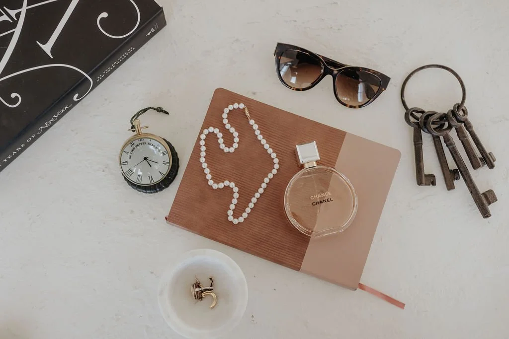 A flat lay of fashionable accessories including sunglasses, a lipstick, a watch, a pearl necklace, a set of vintage keys, a small dish with a pair of earrings, and a Chanel perfume bottle on a white surface.