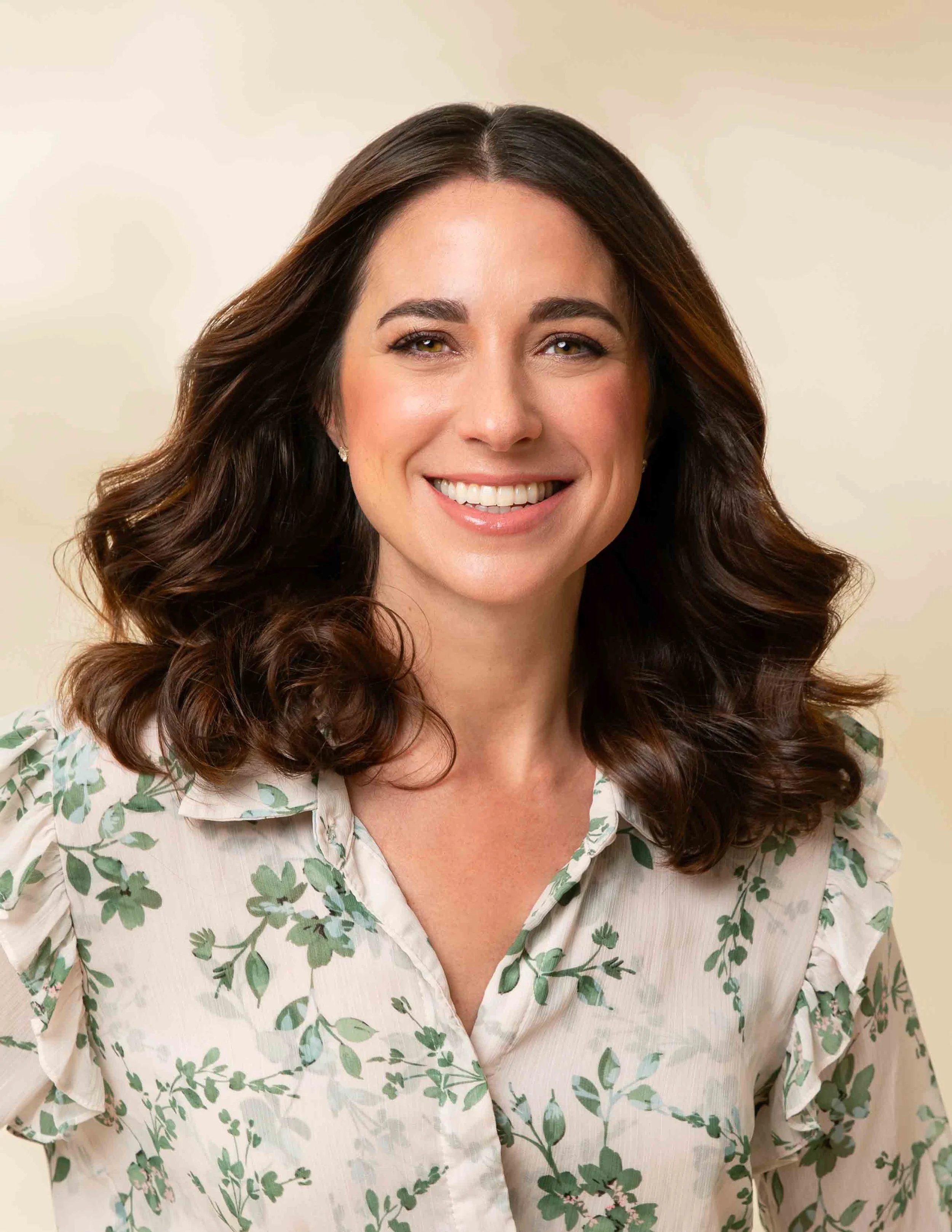 A woman with long, dark, curly hair smiling in front of a beige background, wearing a light-colored blouse with green leafy pattern.