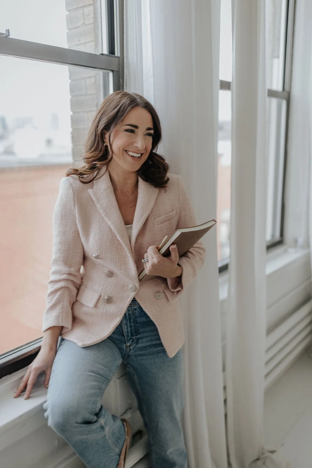 A woman with dark brown hair and light skin, wearing a light pink blazer and jeans, smiling while holding a beige notebook, standing by a window with white curtains, in a room with large windows and white walls.