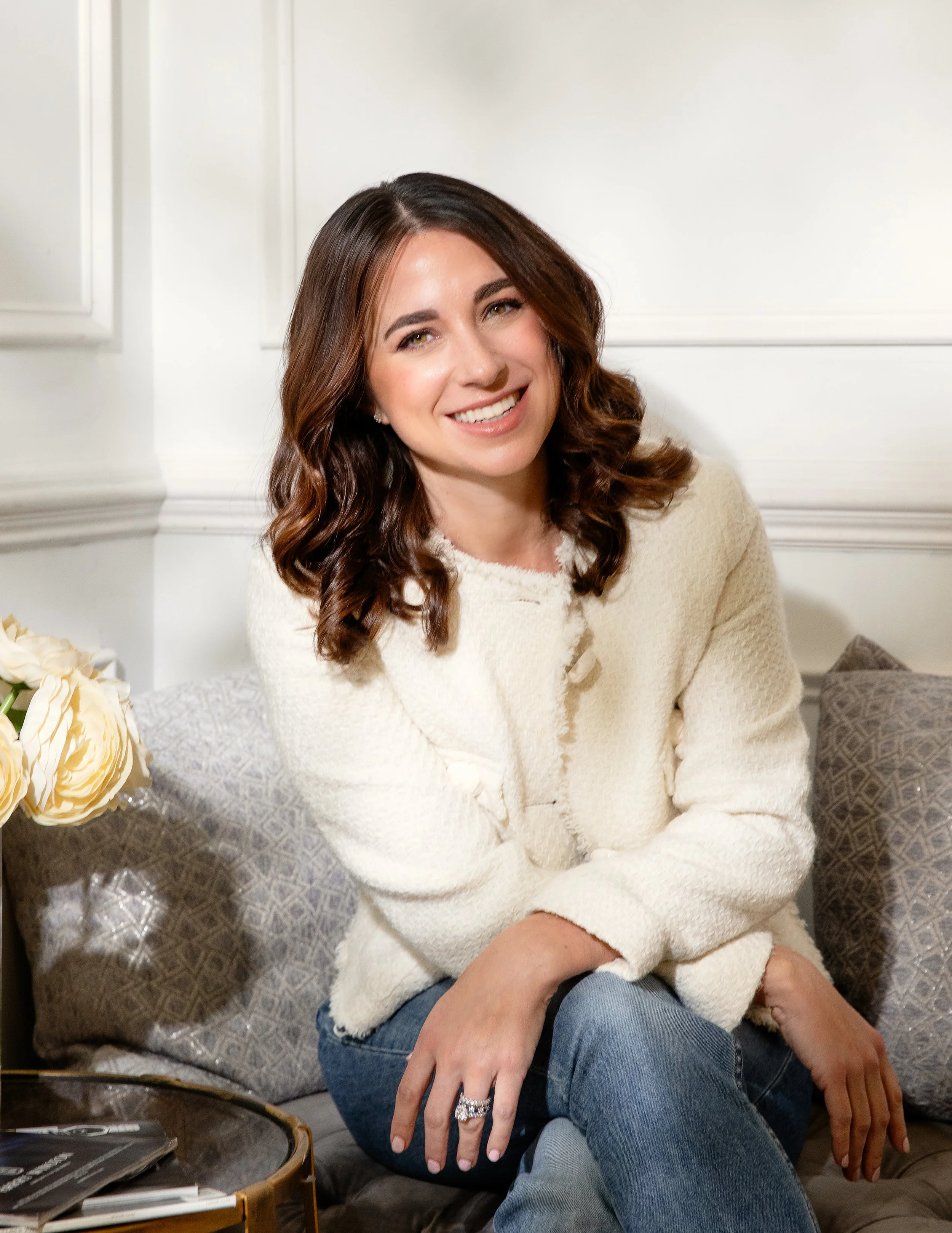 A woman with brown hair, wearing a beige sweater and blue jeans, sitting on a sofa, smiling and looking slightly to the side.